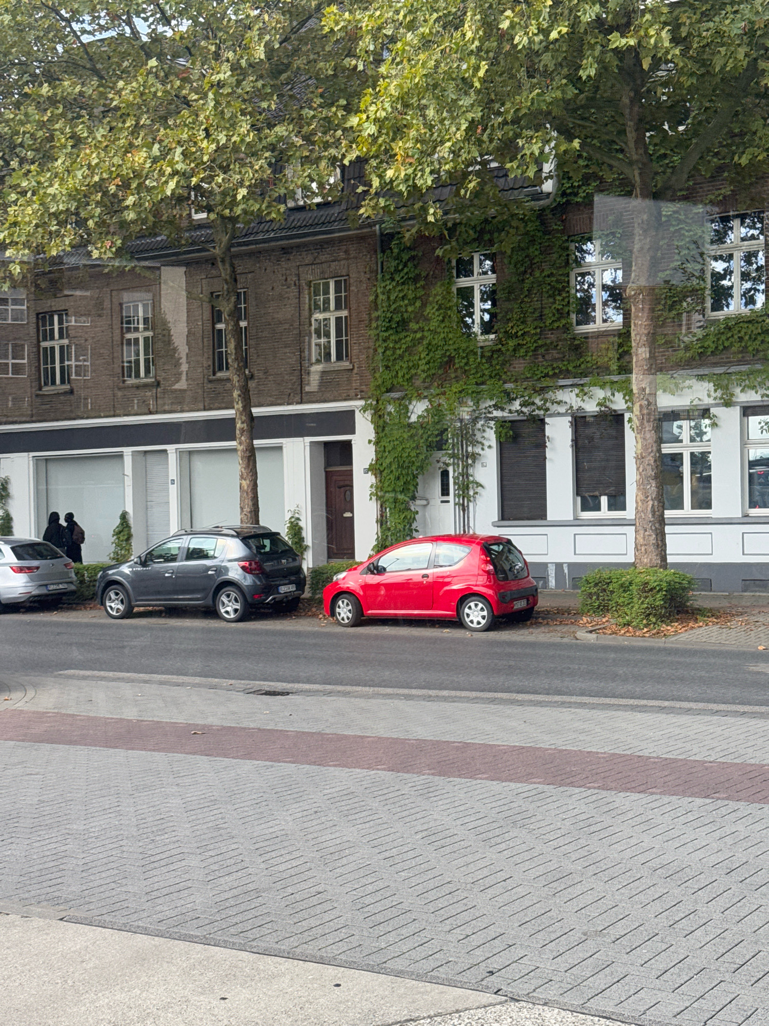 The image depicts a street scene in the city of Cleves, Germany, featuring a building facade with trees and parked cars. The building is made of dark brick and is partially covered in ivy, with several windows and a door visible. In front of the building, a grey car, a red car, and a silver car are parked along the curb. Two individuals, appearing to be women based on their silhouettes and hairstyles, are walking together on the sidewalk in front of the building, near the grey car. The street in the foreground is paved with grey and red bricks, forming a patterned surface. The scene is illuminated by daylight, casting shadows on the street.