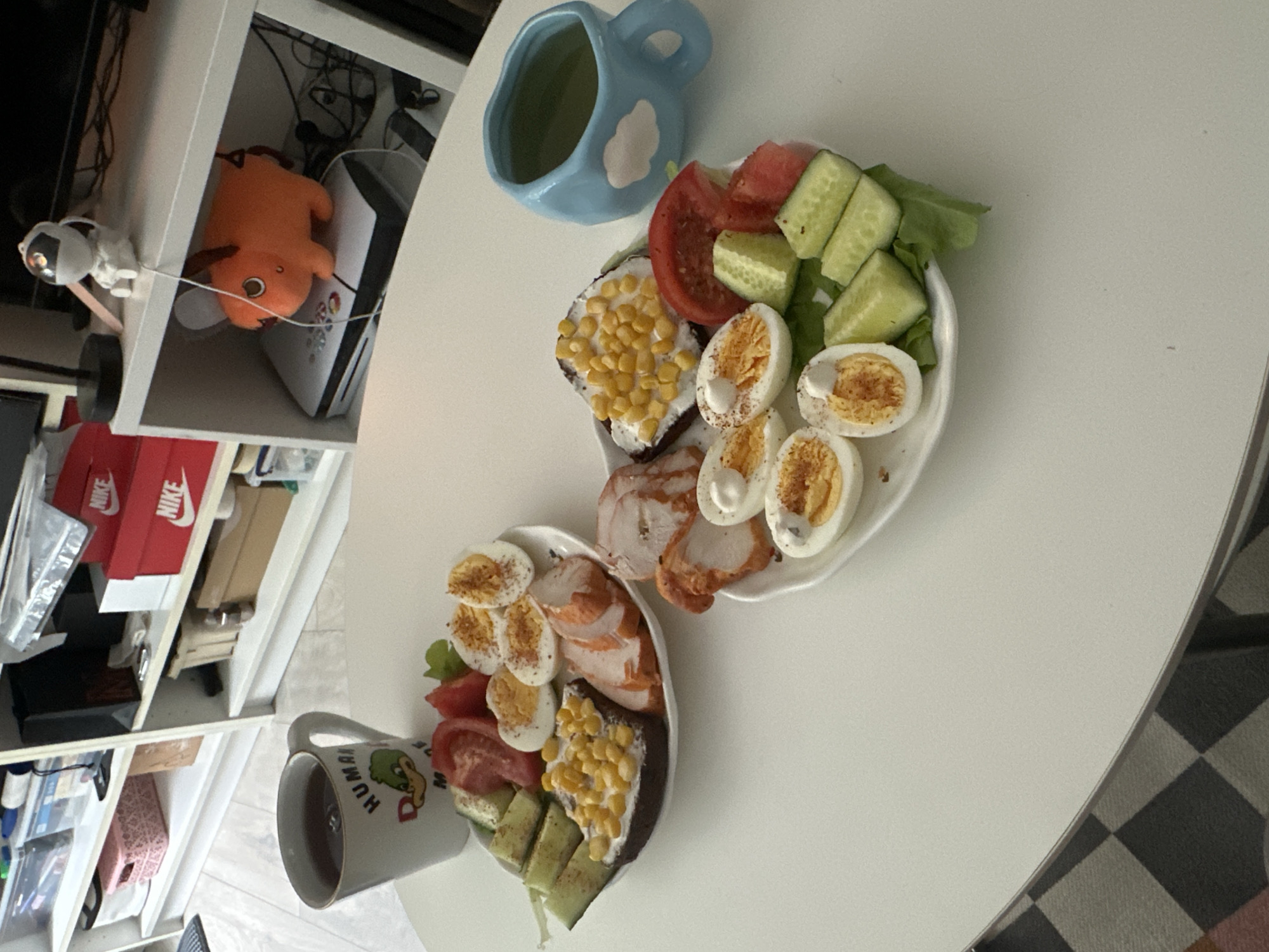The image displays a close-up, overhead shot of two white plates filled with food, set on a white table. The plates contain a breakfast or light meal consisting of sliced hard-boiled eggs, sliced tomatoes, cucumber wedges, lettuce, corn, and sliced cooked chicken. One plate also features what appears to be a slice of dark bread topped with a white spread and corn. To the left of the plates, a white mug with a graphic of a duck and the text "HUMAN MADE" is visible, containing a dark liquid. In the background, a light blue ceramic mug with a cloud design rests on the table. To the left of the table, a white shelving unit is partially visible, with various items including a bright orange plush toy, a white figurine, and red Nike shoeboxes. The lighting suggests daytime. The overall impression is a casual meal setting.