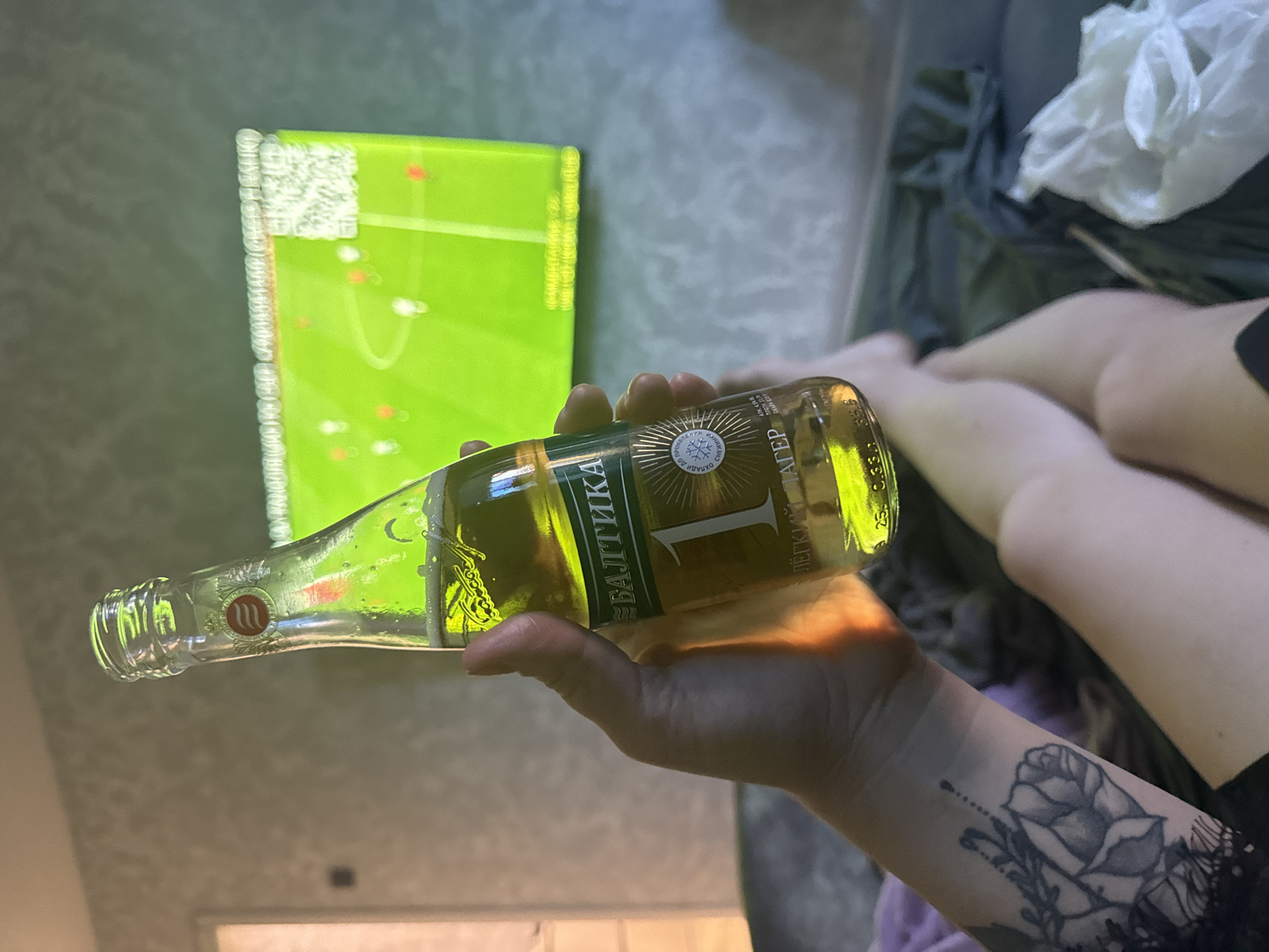 A person is holding a bottle of "Baltika 1 Lager" beer in their right hand. The person has a floral tattoo on their wrist. In the background, there is a television displaying a soccer game. To the right of the person's hand, a portion of a leg and a crumpled plastic bag are visible on what appears to be a couch. The environment suggests an indoor setting, possibly a living room. The location is unknown.