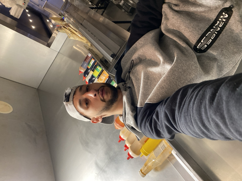 A man with a beard and a baseball cap takes a selfie in what appears to be a commercial kitchen. He wears a grey and blue hoodie. In the background, there's stainless steel equipment, condiment bottles, and various sauces lined up on a counter. Fluorescent lighting is visible in the top left corner. The overall setting suggests the man is in a food service environment.