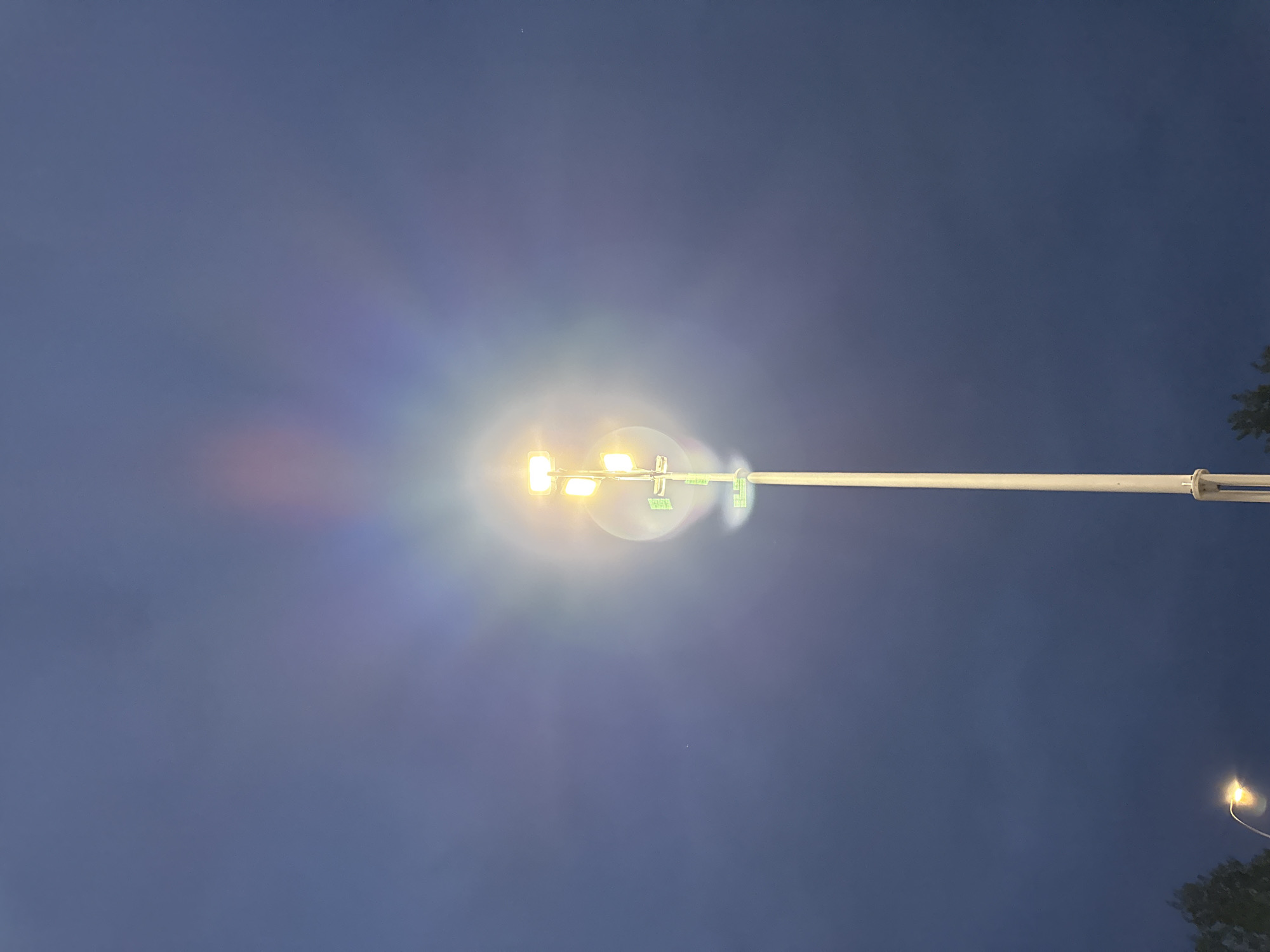 A tall, white street light fixture stands prominently in the center of the frame against a dark blue, cloudy sky. The fixture has multiple rectangular light panels, with the top ones emitting a bright yellow glow. Surrounding the lights, there is a visible halo effect with rainbow-like colors, suggesting a lens flare or atmospheric phenomenon. The pole is segmented, with visible circular mounting points. In the lower left corner, the top of a smaller street light can be seen, also emitting a yellow light. The lower edges of the frame show dark foliage, hinting at trees. The overall scene appears to be an outdoor urban environment at dusk or during low light conditions.