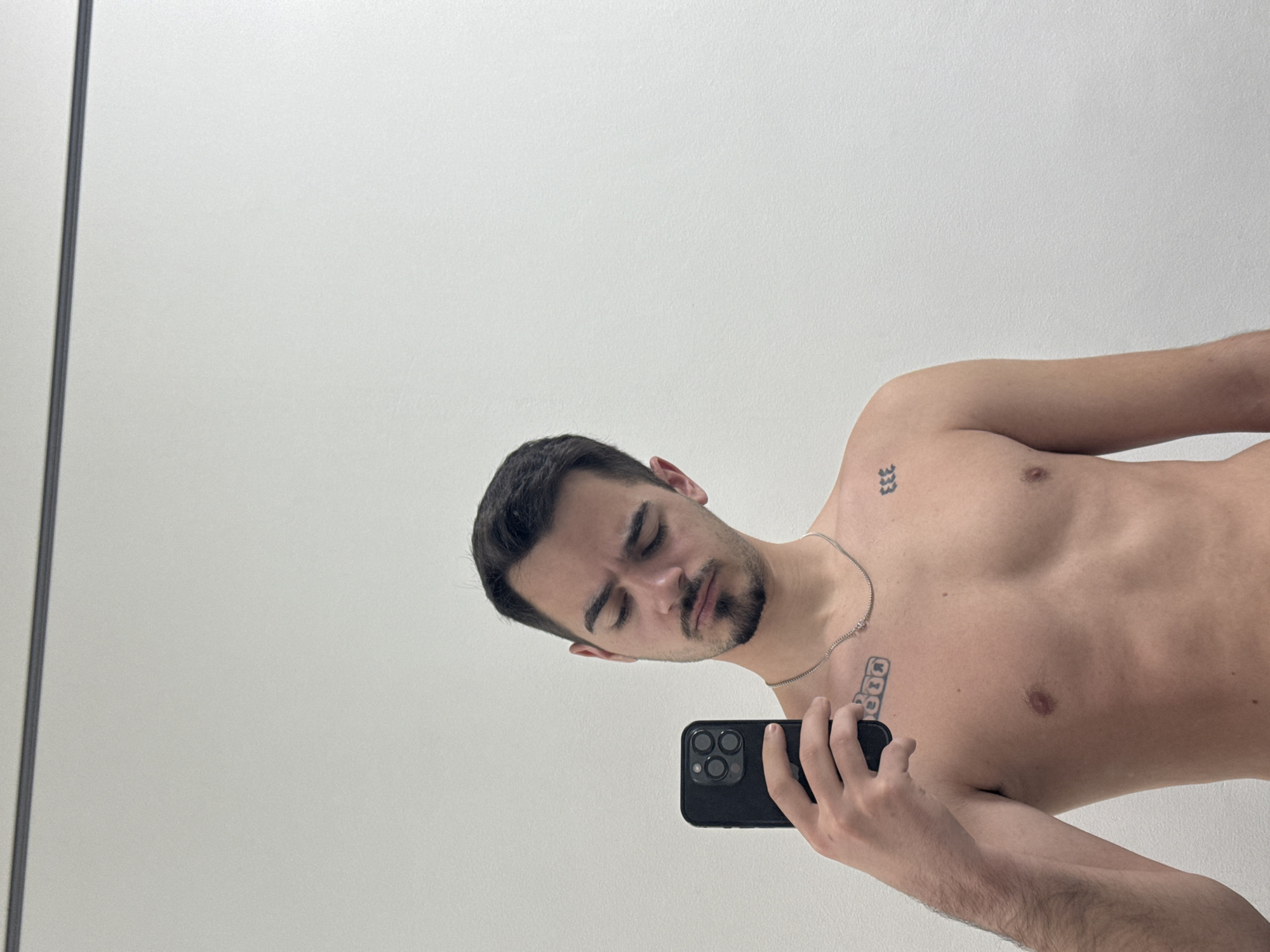 A bare-chested man is posing in front of a mirror while holding a black smartphone in his right hand. He has dark hair and a trimmed beard. Tattoos are visible on his upper chest and shoulder. The background is a plain white wall with a thin dark line suggesting the edge of the mirror frame. The image does not contain information to ascertain the location in the city of Sofia, Bulgaria.