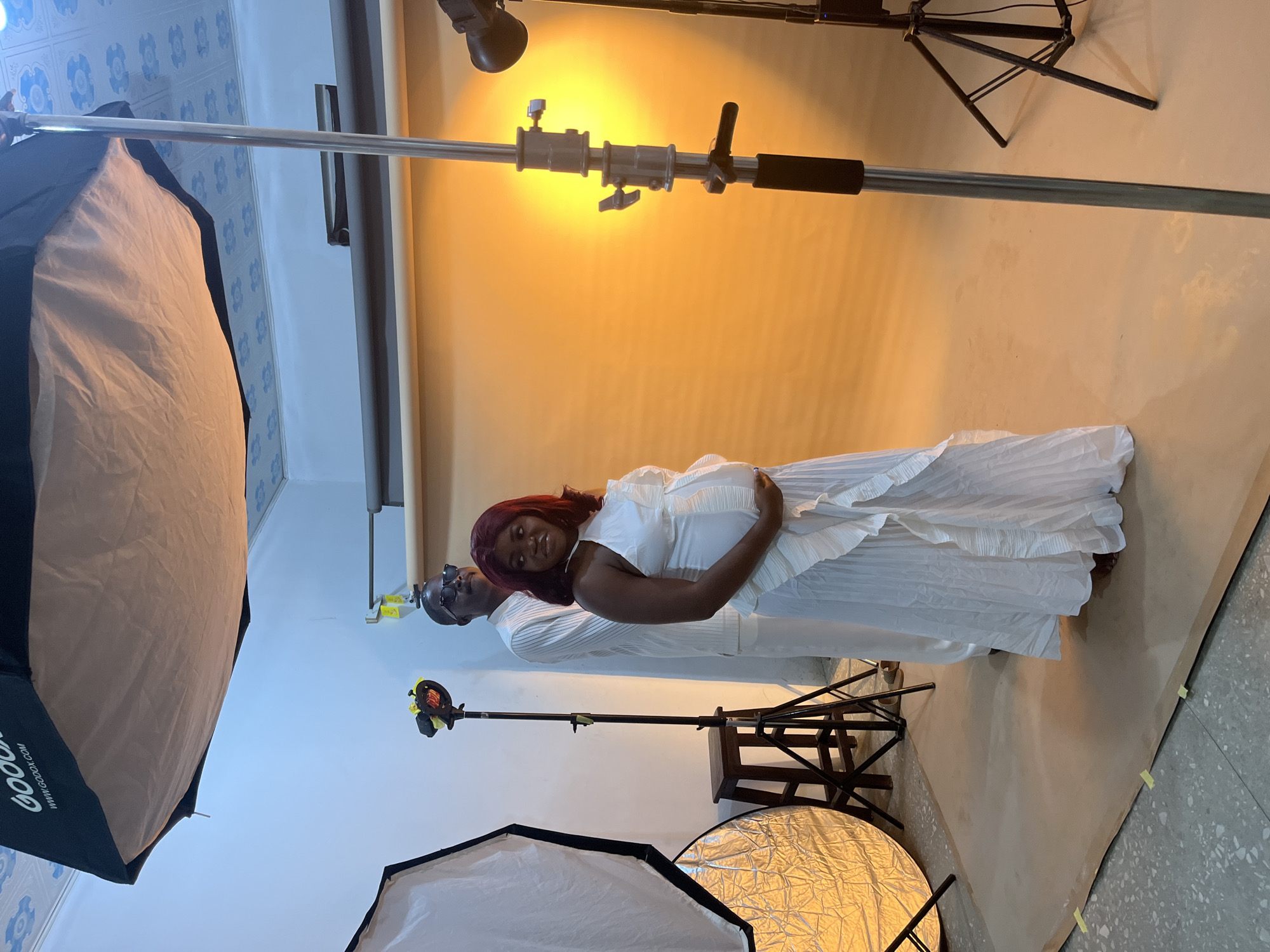 A woman in a white dress is standing in a photography studio, holding her pregnant belly, with a man in a white shirt and glasses standing behind her. The background is a seamless yellow paper backdrop. Several pieces of photography equipment surround the couple, including softboxes, reflectors, and lighting stands. The floor is a gray terrazzo. The location is a photography studio. There is no evidence that the photo was taken in Gura Top, Nigeria.