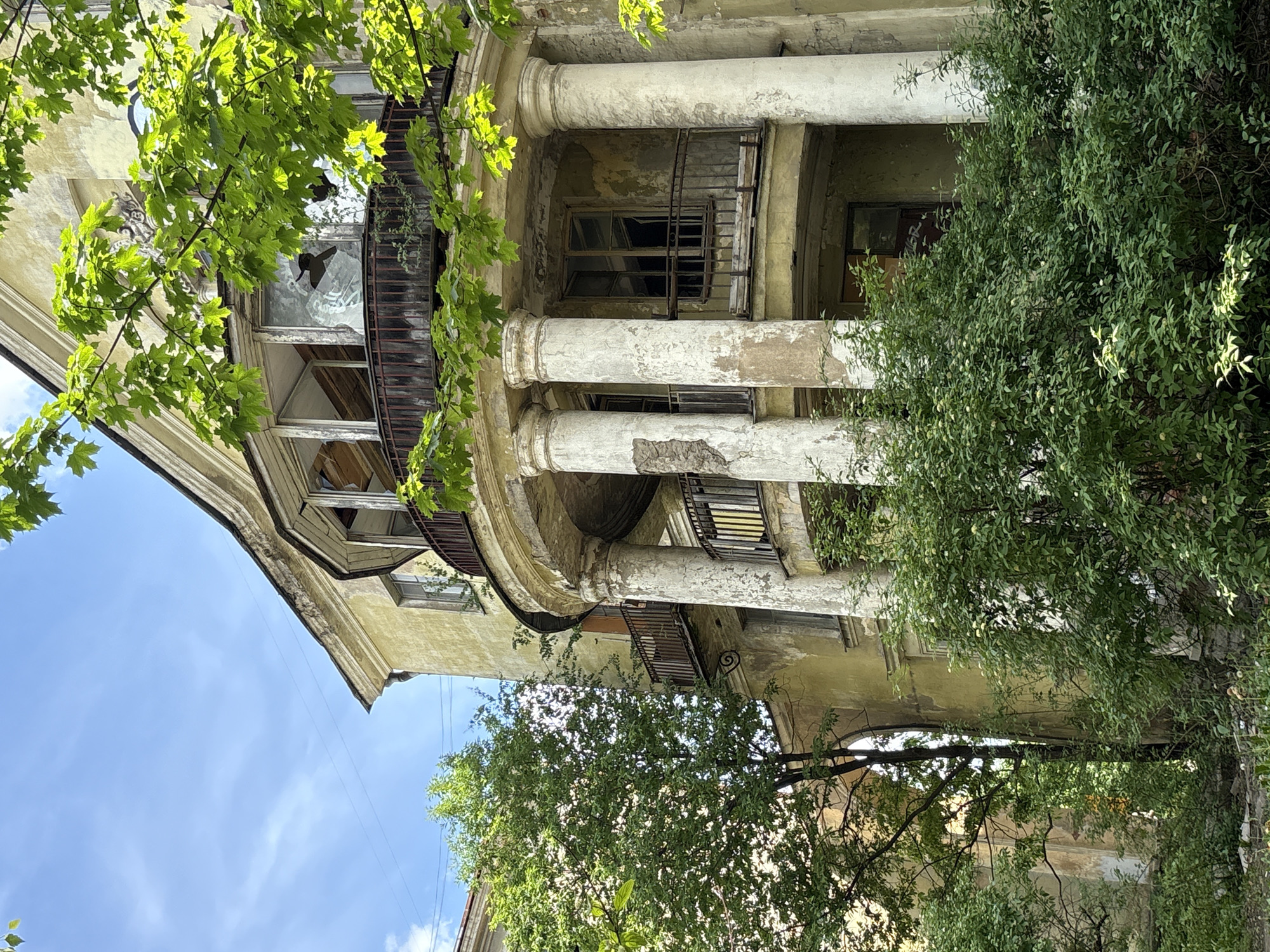 A weathered, pale yellow building with peeling paint and arched windows stands amidst overgrown greenery under a bright, partly cloudy sky. The building features a prominent rounded balcony supported by three fluted columns, showing signs of decay and damage. The balcony railing is rusted, and some panes of glass in the enclosed upper portion of the balcony are broken, revealing the interior. To the left, a lush green tree with dense foliage partially obscures the building's facade. To the right, more dense green bushes and trees grow in front of the building, some with small white flowers. The scene appears to be in an urban setting, possibly St. Petersburg, Russia, given the architectural style and the presence of greenery juxtaposed with an aged building. There are no people visible in the image. The overall impression is one of neglect and the encroaching power of nature on abandoned structures.