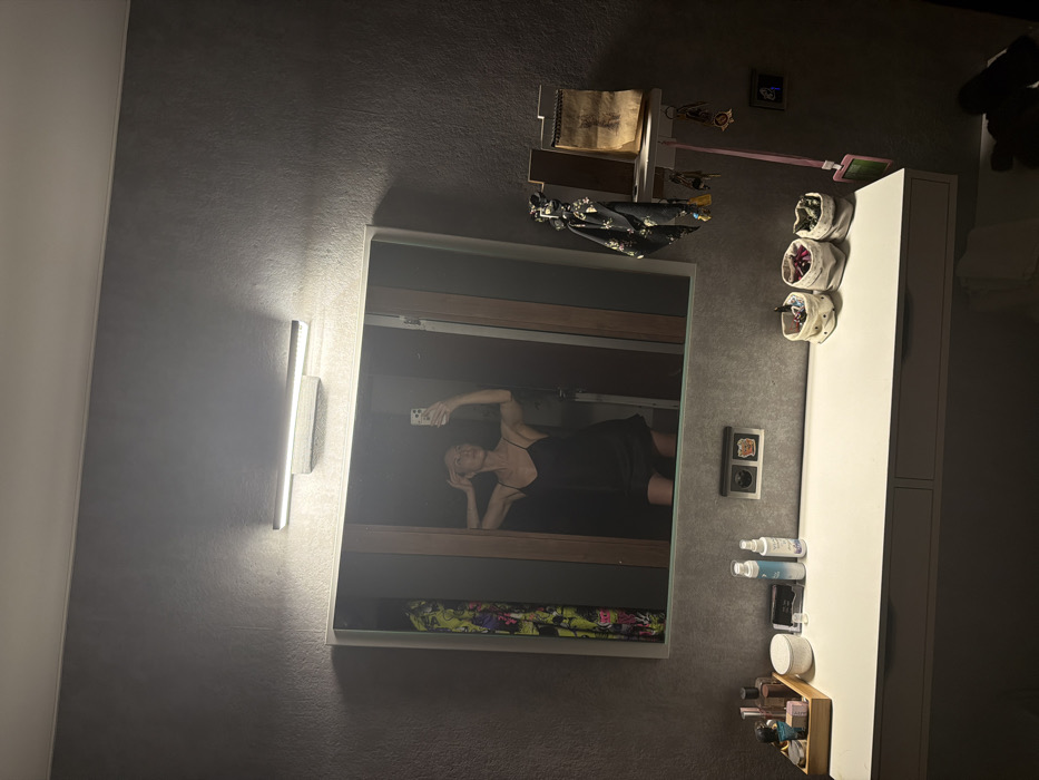 A bathroom scene features a mirror reflecting a person taking a selfie. The individual wears a dark dress and reclines on a bed-like surface within the mirrored reflection. To the right of the mirror, a white vanity with cylindrical storage containers is visible. Below, toiletries are arranged. A light fixture adorns the left wall beside the mirror, while a shelf with trinkets is mounted above it. The overall ambiance is dimly lit.