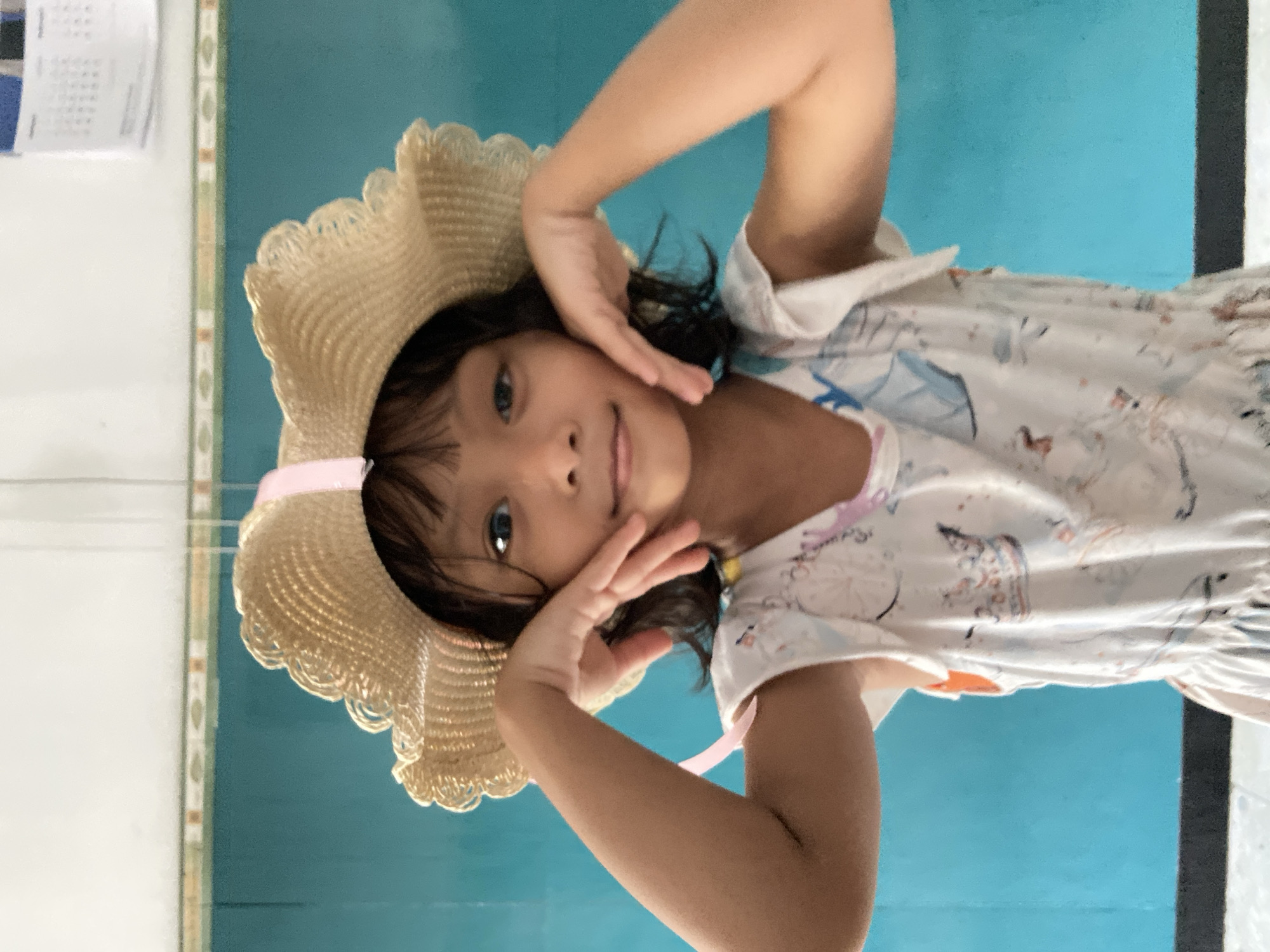 A young Asian girl with short dark hair and a straw hat with a pink ribbon is posing for a photograph. She is wearing a light-colored dress with a whimsical pattern. She is positioned indoors, likely in Gelam, Indonesia, against a wall painted in shades of blue and white. Her hands are framing her face, and she is looking directly at the camera.