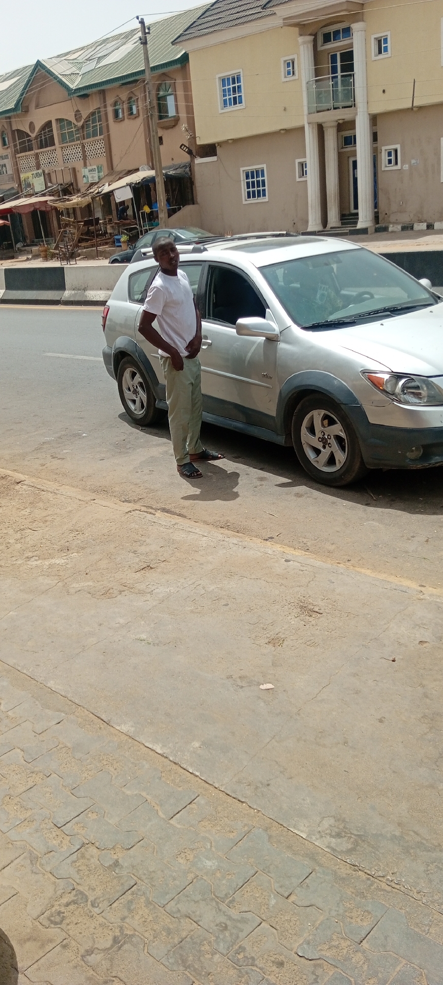 The image shows a man standing beside a silver car on a street in Fagge D2, Nigeria. The man is dark-skinned, wearing a white shirt and light-colored trousers with sandals. He is looking towards the camera with a slight smile. The car is a compact SUV with a sunroof. The background includes a multi-story building with storefronts and a residential building with white columns. The ground is a combination of concrete sidewalk and paved road. It is daytime, with sunny conditions.