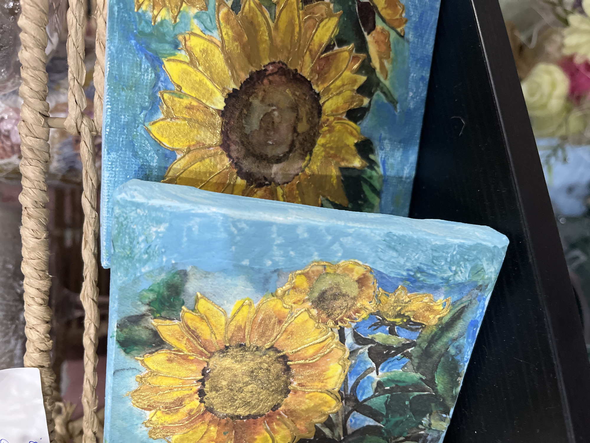 The image shows two small paintings of sunflowers, each on a square canvas with a light blue background. The sunflower in the top painting is more prominent and closer to the viewer, with a dark brown center. The bottom painting shows a cluster of sunflowers with golden yellow petals and green leaves. The paintings appear to be displayed indoors, possibly in a store or gallery. A woven, rope-like material is visible to the left of the paintings. To the right, a black frame suggests the paintings may be on display in a shelving unit. The location is unknown.