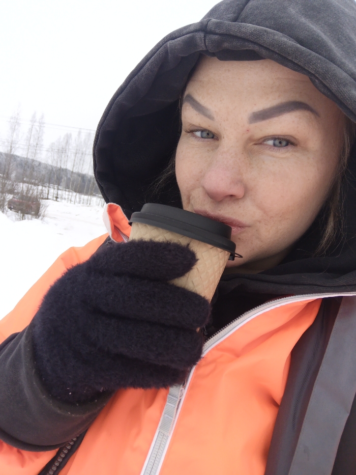 A close-up outdoor portrait depicts a female individual in the city of Svetloe, Russia, during winter. The subject, with light eyes and facial freckles, wears a dark hooded garment under an orange high-visibility jacket featuring grey reflective stripes. Her left hand, covered by a dark knitted glove, holds a light brown, corrugated disposable cup with a black lid, positioned near her mouth as if consuming its contents. The background reveals a snow-covered landscape with bare deciduous trees, distant hills, and utility lines under an overcast sky. A red, indistinct object is partially visible in the far left background.