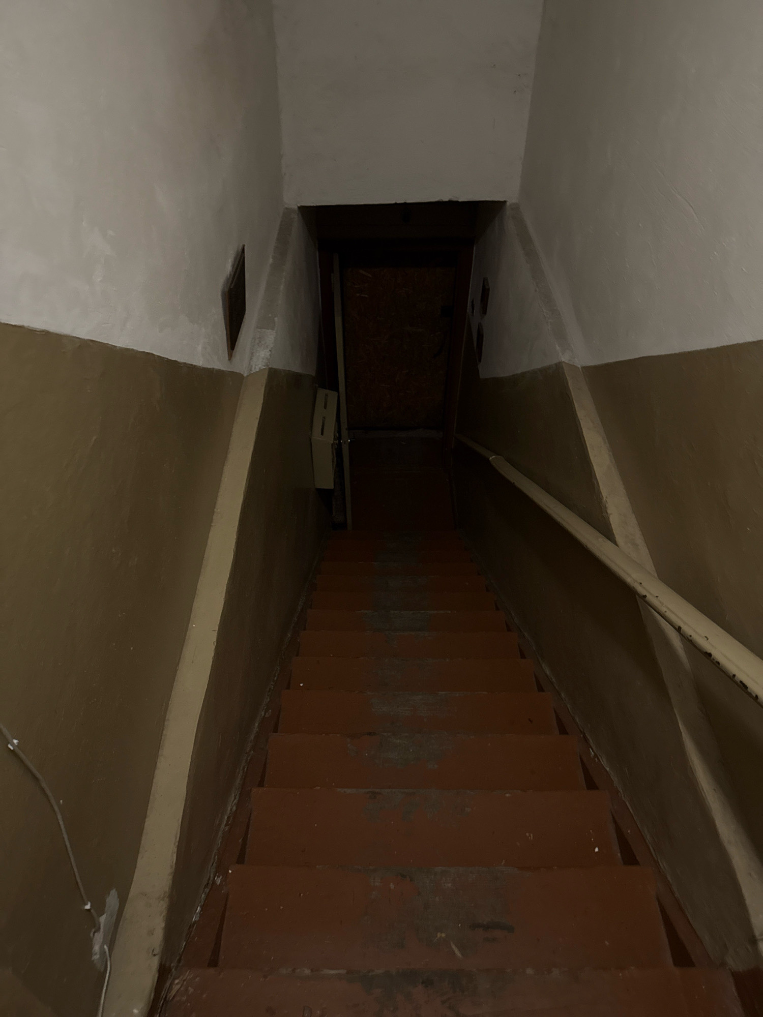 The image shows a dimly lit, narrow staircase in what appears to be a residential building in Kaliningrad, Russia. The walls are painted in two distinct tones: a lighter cream color on the upper portion and a darker, earthy brown on the lower portion. A light-colored, somewhat worn handrail runs along the right side of the staircase. The stairs themselves are painted a dark red or rust color and show signs of wear and tear. At the top of the staircase, a wooden door is visible, appearing to lead to another area or room. A light-colored electrical box or intercom is mounted on the wall to the left of the door. The overall atmosphere of the image is somewhat somber and utilitarian.