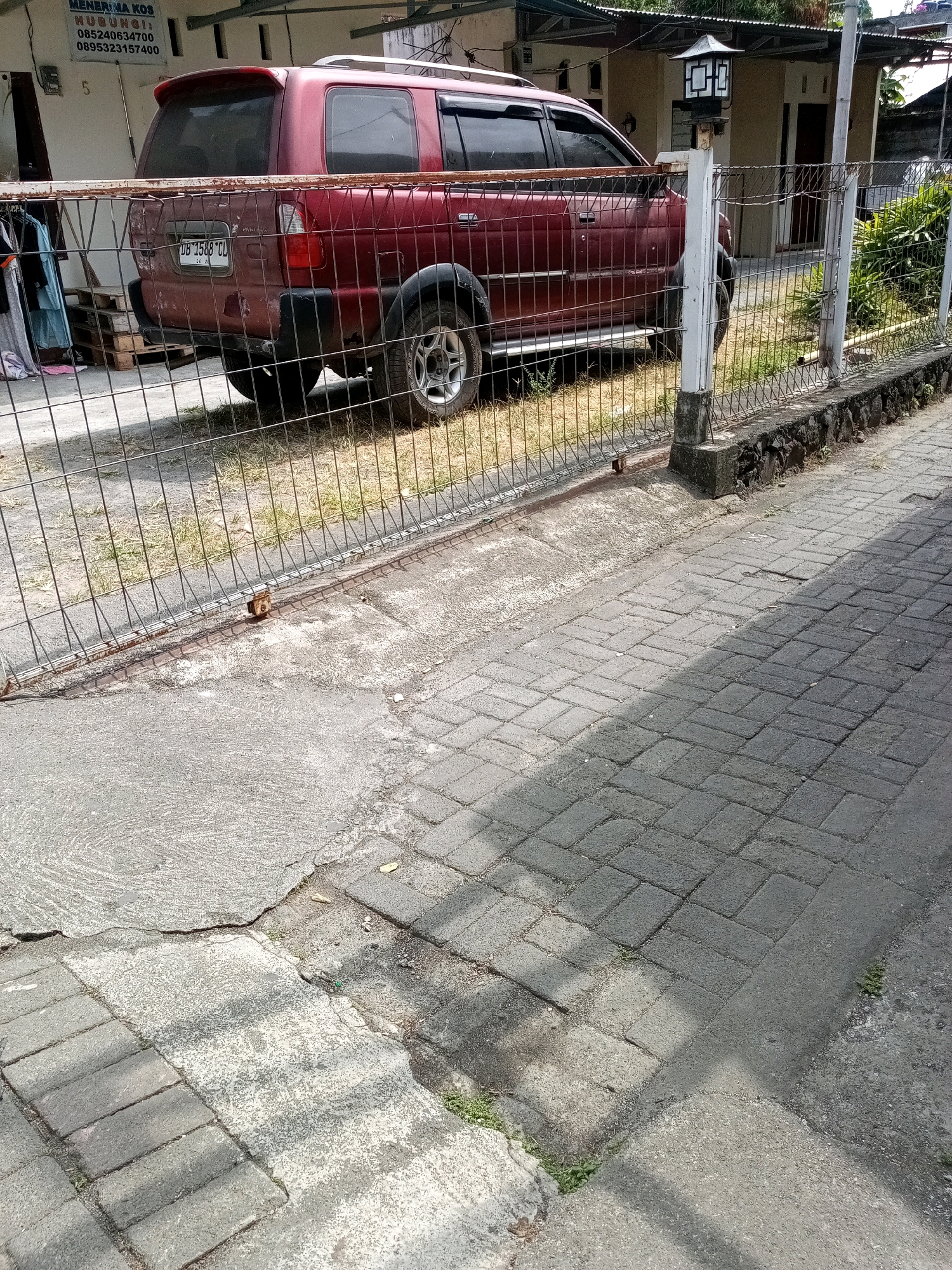 A maroon SUV is parked behind a metal fence. The vehicle has tinted windows and a spoiler on the rear. The license plate is visible and reads "DB 1568 CU". The SUV is parked on a grassy area next to a paved walkway. The walkway is made of interlocking bricks and concrete. There is a house with a light fixture on a pole in the background. The scene appears to be in a residential area. The image was taken in Bitung, Indonesia.