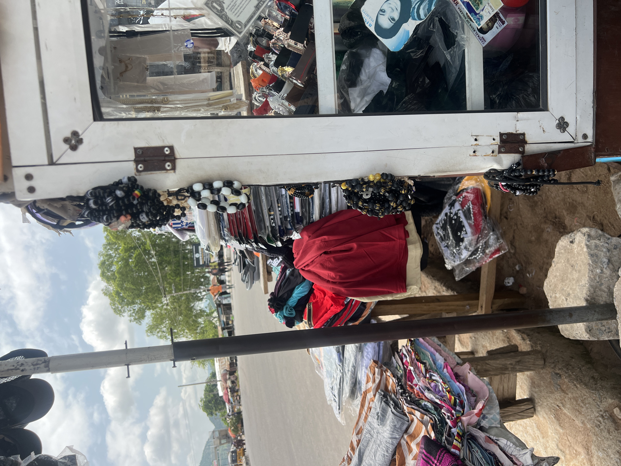 This image depicts an outdoor market scene in Howayi, Nigeria. The foreground is dominated by a display of various items, including colorful folded clothing, possibly t-shirts or wraps, and several strands of beaded bracelets. The bracelets are made of black and white beads, some with yellow accents. A red garment, possibly a shirt or jacket, is prominently displayed among the folded clothes.
Behind the merchandise, a white structure with a glass display case is visible. Inside the case, more items are displayed, including what appear to be watches, jewelry, and possibly some currency or documents, one of which shows a portrait and text. The glass is slightly smudged, and reflections suggest an outdoor setting. The structure seems to be part of a market stall.
The background reveals a street with a few vehicles and people walking, suggesting a moderately busy area. Green trees and a partly cloudy sky are visible, indicating daylight. The overall impression is of a bustling local market with goods laid out for sale. The ground appears to be a mix of dirt and possibly some gravel. The orientation of the image is rotated, with the market stall and street appearing vertically aligned.