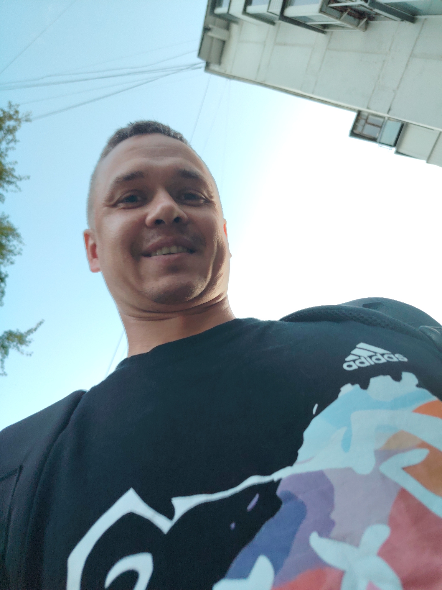A man is taking a selfie from a low angle, looking up at the camera. He has short, dark hair and is smiling. He is wearing a black t-shirt with a colorful graphic design on it, and the Adidas logo is visible on the upper right side of the shirt. Behind him, a modern building with many windows can be seen, and the sky is clear and blue. The image appears to have been taken outdoors, likely in an urban setting in the city of Chaikovsky, Russia.