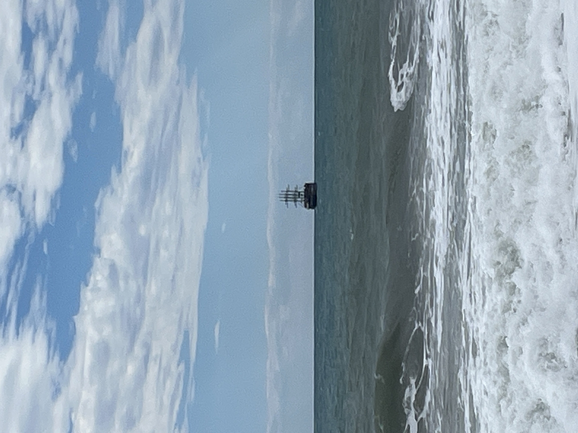 The image shows a seascape, likely taken from the shore in Alanya, Turkey. A tall sailing ship is visible on the horizon, relatively far from the coastline. The sky is partly cloudy, with visible blue patches. The sea closer to the shore is choppy, with white waves breaking. The photo is taken at an unusual angle, rotated 90 degrees.