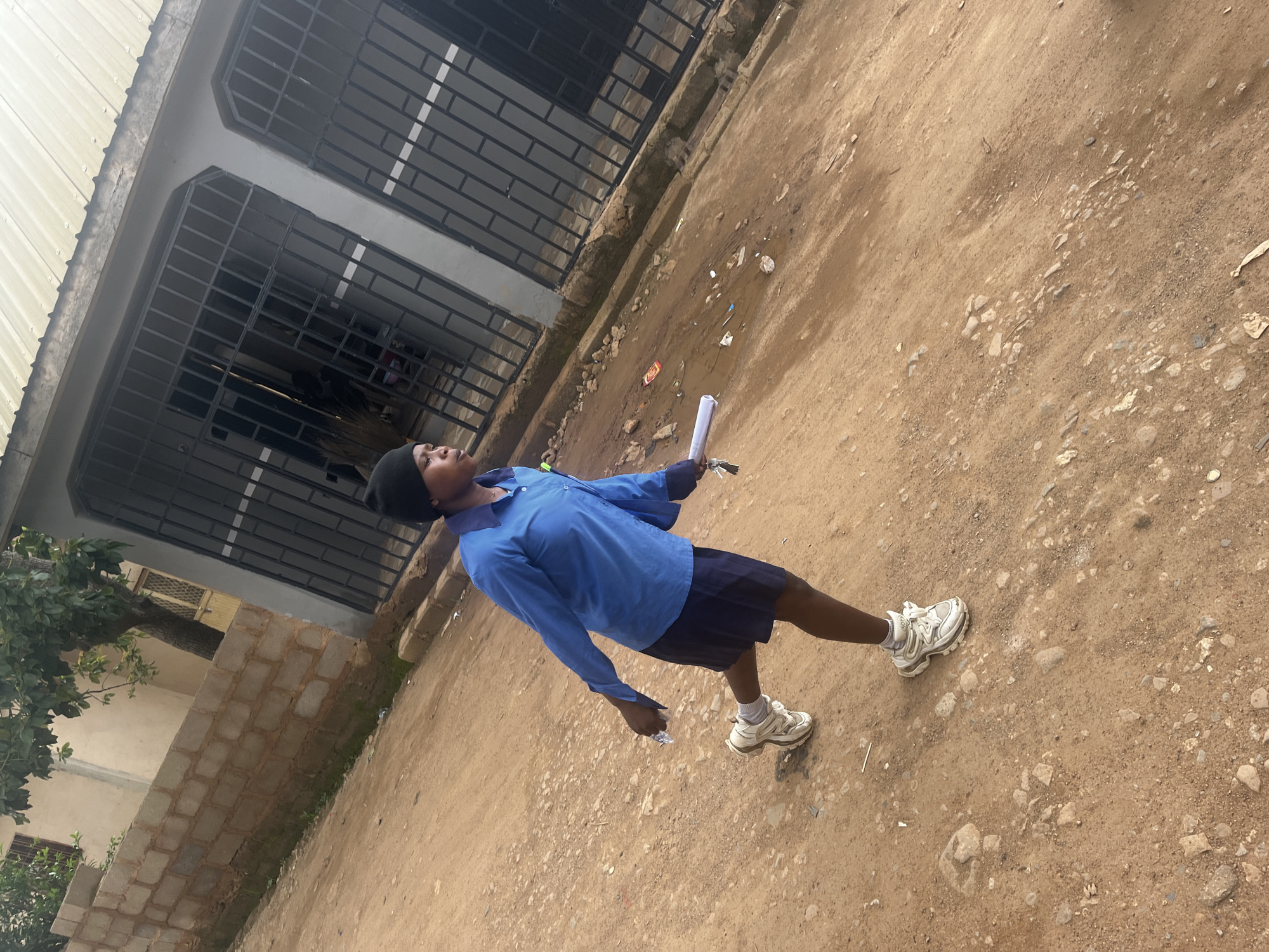 In Gura Top, Nigeria, a young person wearing a blue shirt, dark skirt, and a black beanie walks on a dirt path. They are carrying a rolled-up white paper and keys in their hand. The background shows a building with barred windows and a corrugated metal roof. The ground is covered in dirt and scattered debris.