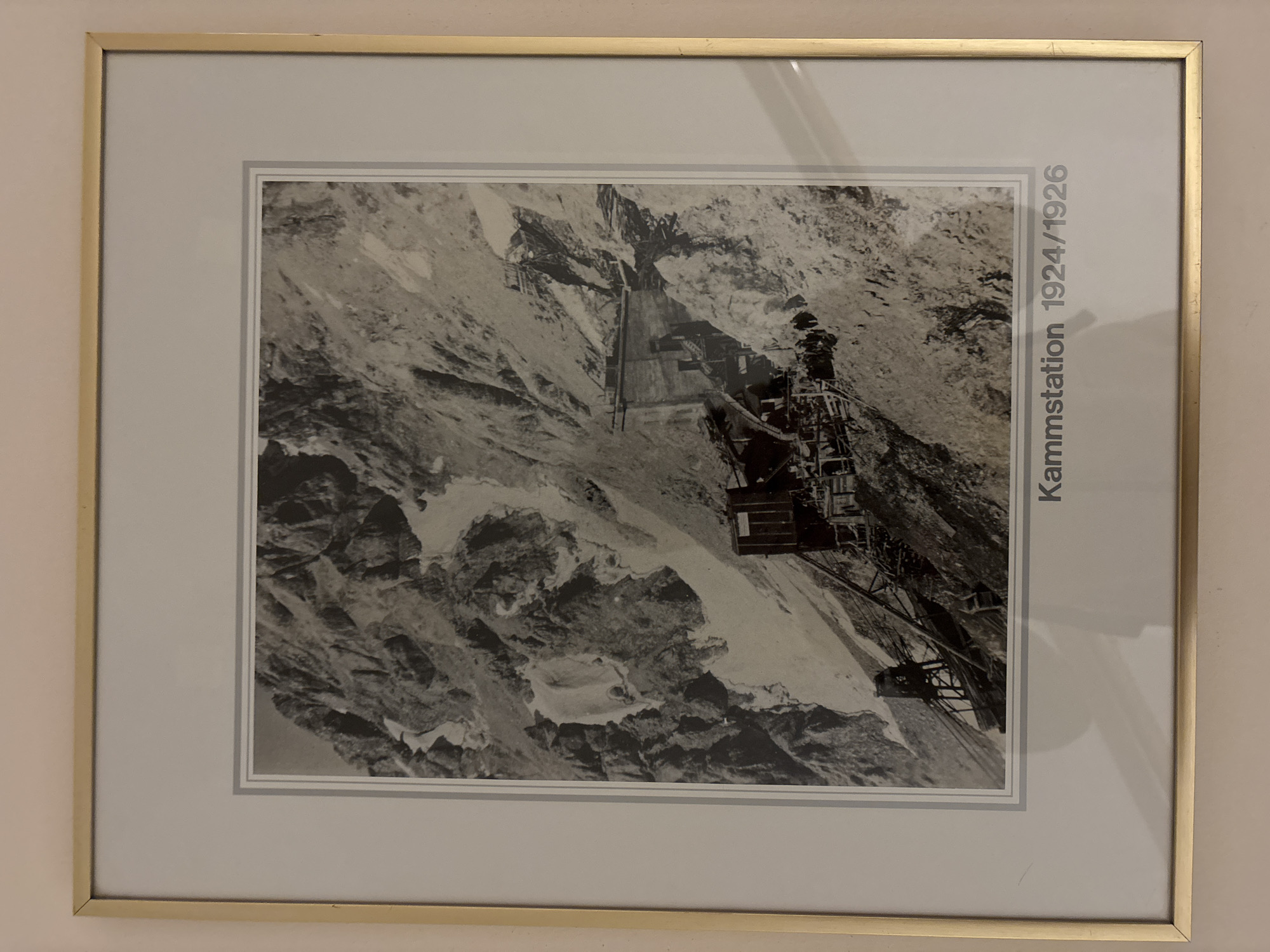 This is a framed black and white photograph depicting a mountain landscape with structures and people. The text at the bottom of the photograph reads "Kammstation 1924/1926," indicating it is a view of the Kammstation (summit station) from the years 1924-1926. The image shows a steep, rocky mountainside with patches of snow. Several wooden structures and what appear to be construction elements, possibly part of a cable car system or building, are visible on the slope. A group of people, small in number and scale, are gathered near some of the structures, suggesting human activity or construction work. The overall scene is rugged and mountainous, with evidence of human intervention in the form of the built structures. The photograph is matted with a light-colored border and framed with a thin gold-colored frame. The location is identified as the Kammstation in Ehrwald, Austria.