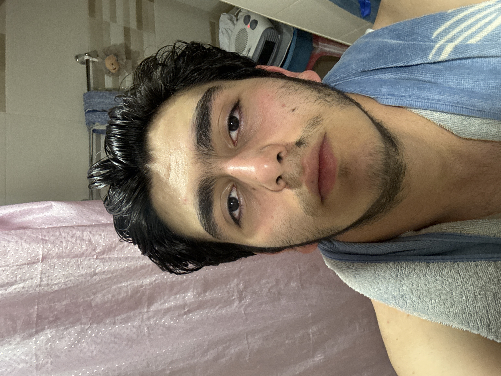A young man with dark hair, dark eyes, and stubble poses for a close-up shot in what appears to be a bathroom. He has a light-colored towel draped around his neck. The bathroom features beige and white patterned tiles and a pink shower curtain. A radiator and a small stuffed animal are visible in the background. The location is likely Yerevan, Armenia.