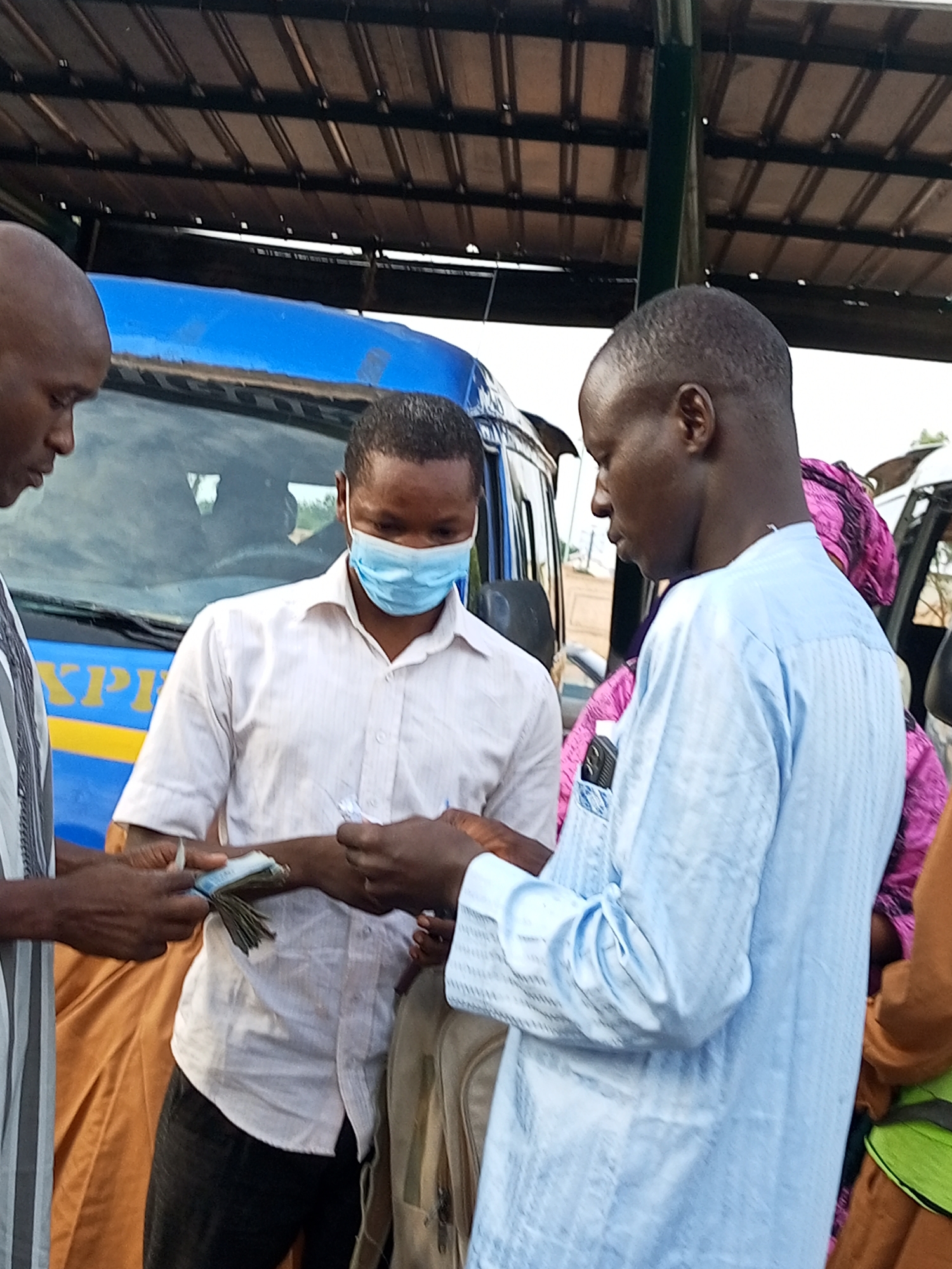 In Gombe, Nigeria, three individuals are observed in what appears to be an outdoor transaction. The central figure, a man wearing a light blue face mask and a white collared shirt, is holding a wad of Nigerian currency, seemingly in the process of handing it to another man on his right. This second man is dressed in a light blue traditional garment. To the left of the central figure, a third man, whose head is visible in profile, is also engaged in the interaction. Behind them, a blue bus with yellow lettering that reads "KPRI" is parked. The scene takes place under a covered structure with a corrugated metal roof, suggesting a market or bus station environment.