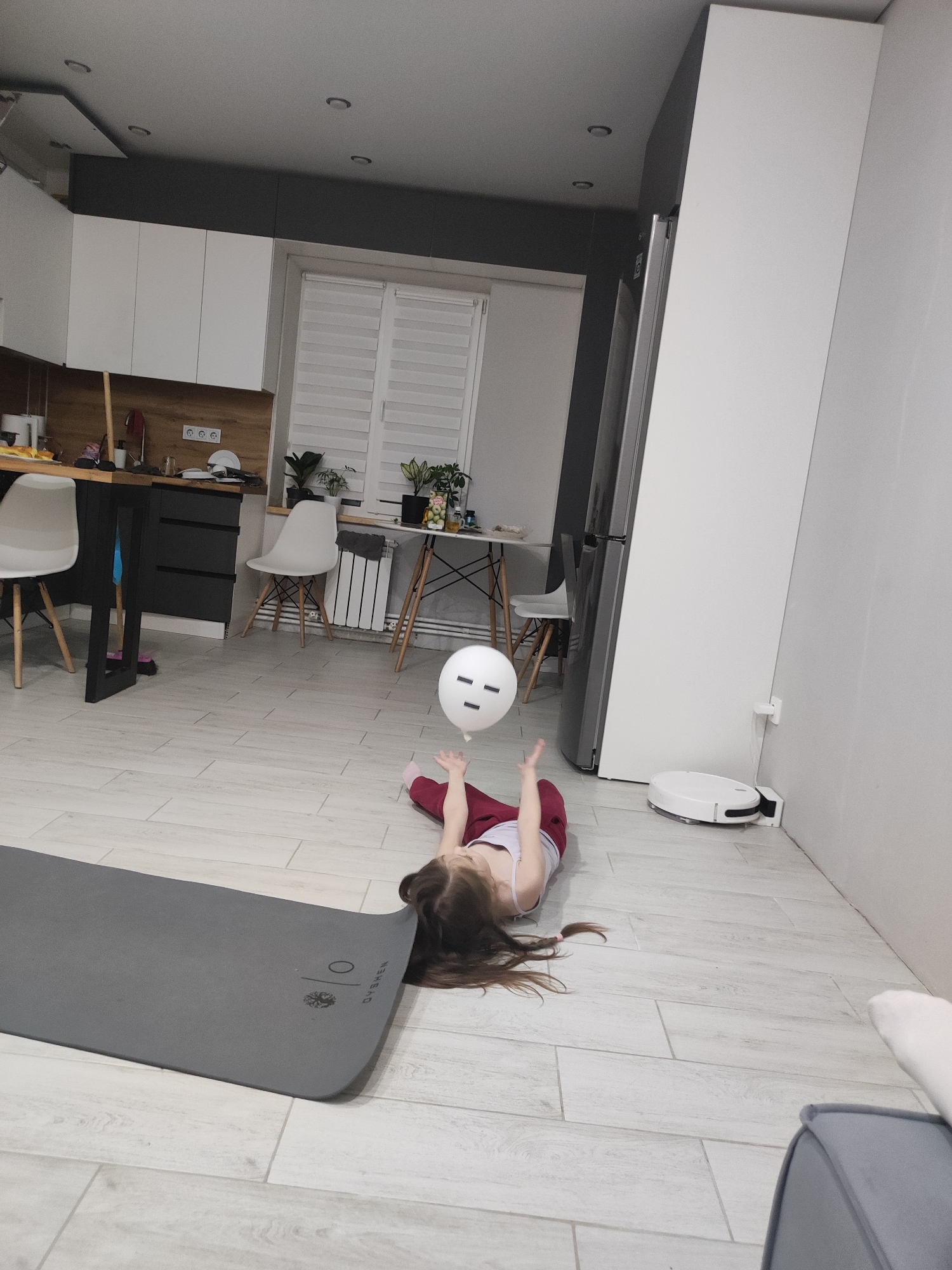 This indoor photograph captures a modern apartment's open-plan kitchen and living area in Votkinsk, Russia. A young child with long brown hair, wearing a white top and maroon pants, lies on their back on a light-colored tiled floor, reaching playfully towards a white balloon with a simple drawn black face (two dots for eyes and a line for a mouth) floating above them.
Adjacent to the child is a gray yoga mat, bearing the visible text "DYSHEN" along with a circular tree-like logo and a "1/0" symbol. The kitchen area features white upper cabinets, dark gray lower cabinets, and a warm wooden countertop. A kitchen island with a dark base and white chairs with wooden legs is visible. A window with white horizontal blinds provides natural light, and a radiator is positioned underneath it, next to a small table with potted plants.
Further into the room, a large gray refrigerator stands next to a tall white cabinet. A white robotic vacuum cleaner is docked and charging against the wall. Recessed spotlights are visible in the ceiling. The overall decor is clean and contemporary, utilizing a palette of white, gray, and natural wood tones. The scene suggests a relaxed and playful atmosphere, likely during the day or early evening.