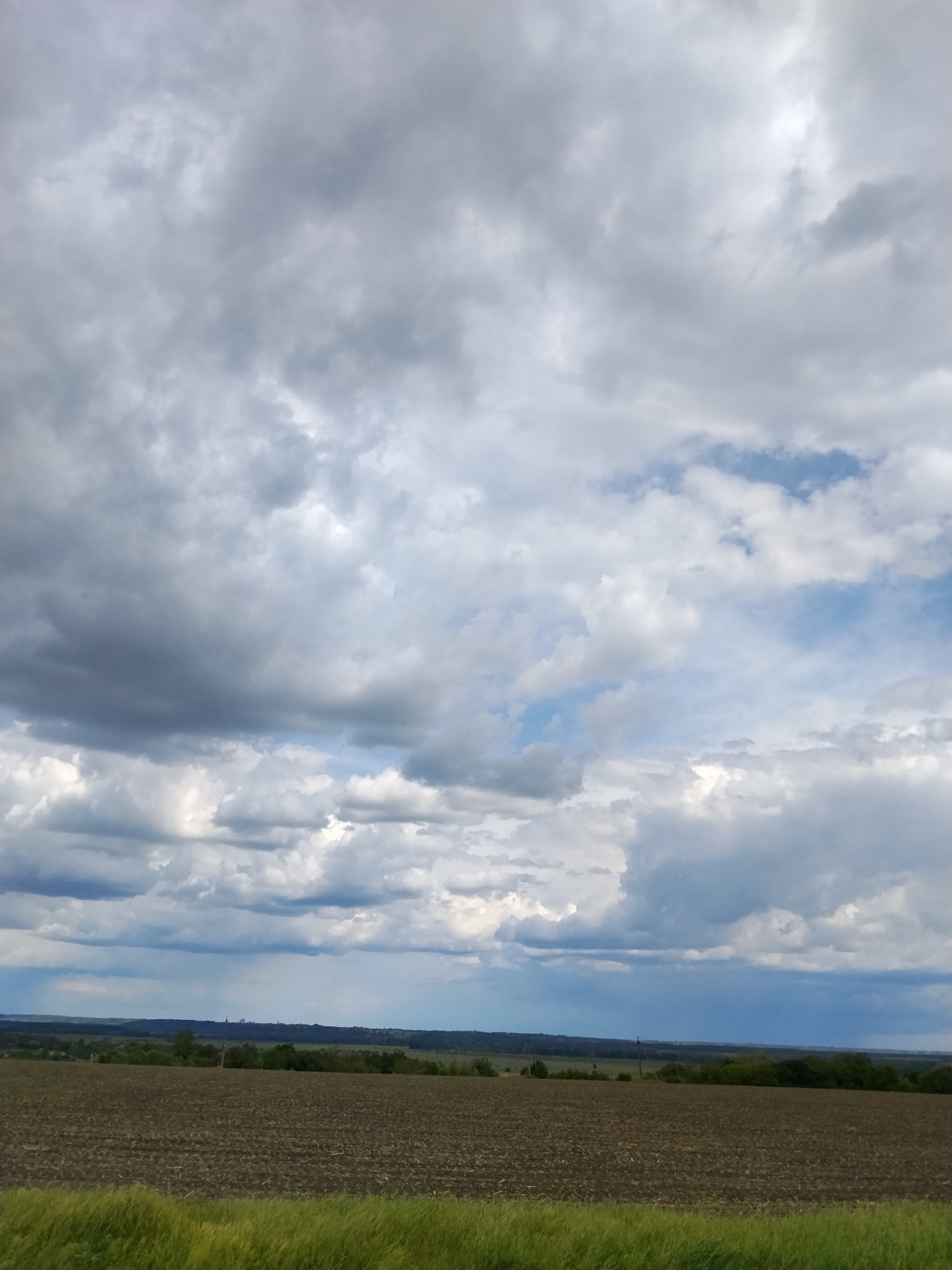 The image shows a rural scene in Ustyvytsia, Ukraine. In the foreground, there's a field of tilled brown soil with a strip of green grass along the bottom edge. A line of trees and vegetation marks the horizon, with a visible drop in the landscape beyond. The sky is dominated by a mix of dark gray storm clouds and patches of light blue. The clouds appear layered, suggesting varying altitudes and weather conditions.