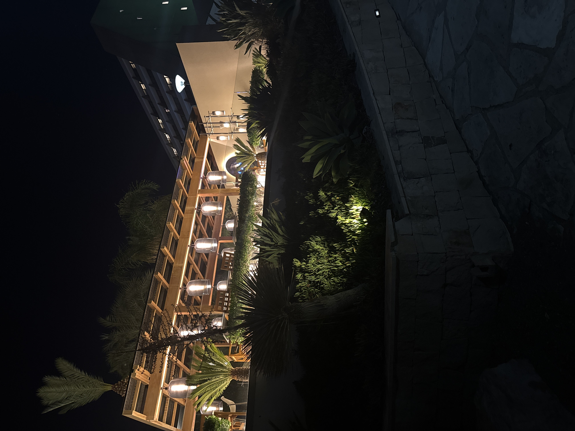The image shows a dimly lit outdoor dining area at night, likely in Agios Tychona Community, Cyprus. A covered, open-air structure with a wooden roof is illuminated by a series of hanging glass pendant lights. Palm trees and other lush greenery surround the area, with some plants highlighted by spotlights. In the background, a multi-story building, possibly a hotel or resort, is visible with its windows lit. The ground in the foreground is paved with irregular stone tiles. There are no people or animals visible in the image.