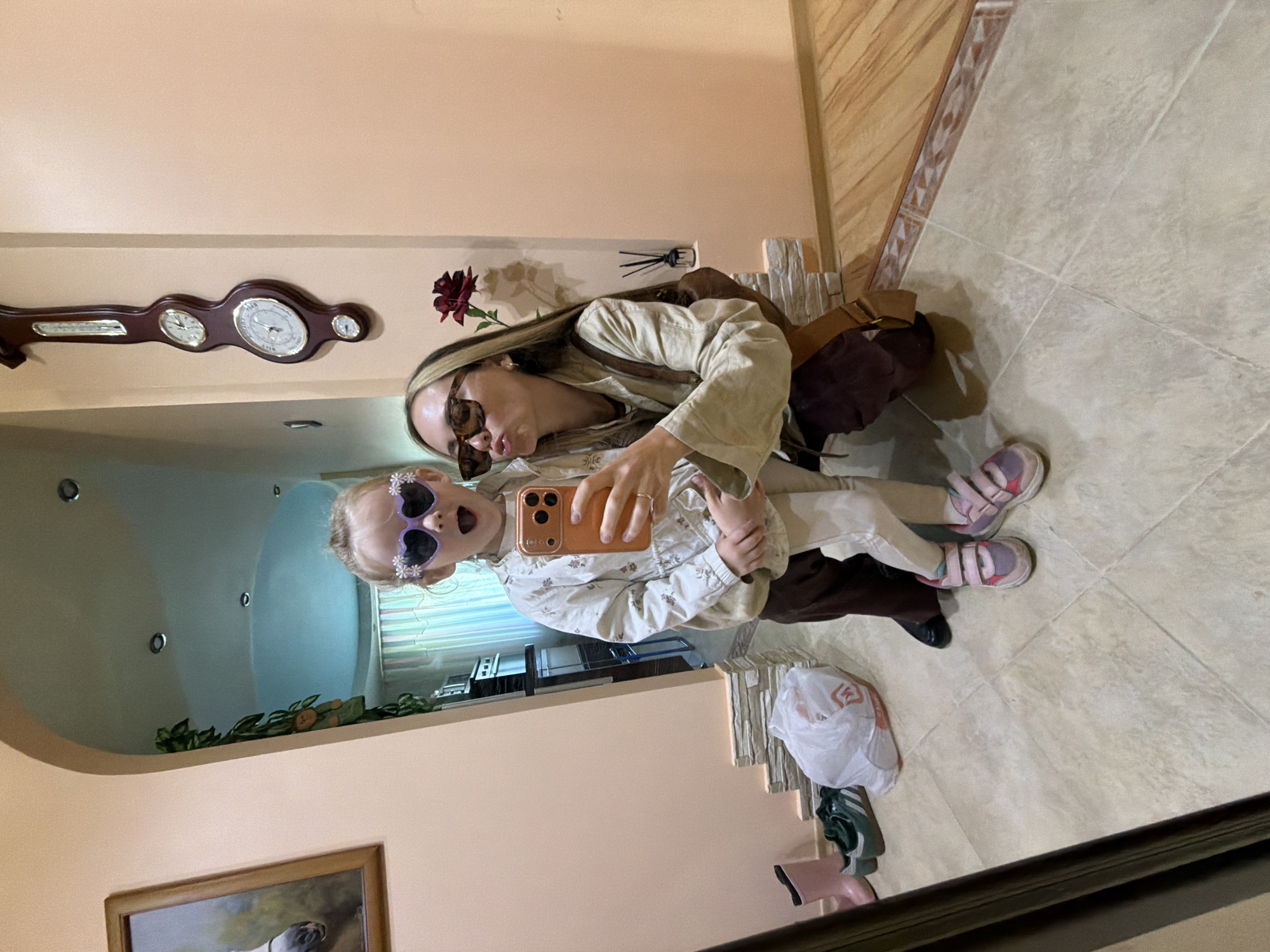 A mirror selfie shows a woman and a young child in what appears to be an indoor setting. The woman is wearing sunglasses and a light-colored jacket, holding a mobile phone, and appears to be taking the picture. The child, also wearing sunglasses in a heart shape with daisy decorations, is being held or posed with by the woman. They both have an excited appearance. The background, reflected in the mirror, includes a wall clock with barometer on the wall, wall decor, as well as items on the floor like shoes, a bag, and plastic bag, all indicative of a domestic environment. Based on additional details, this photo may have been taken in Kamyshin, Russia.