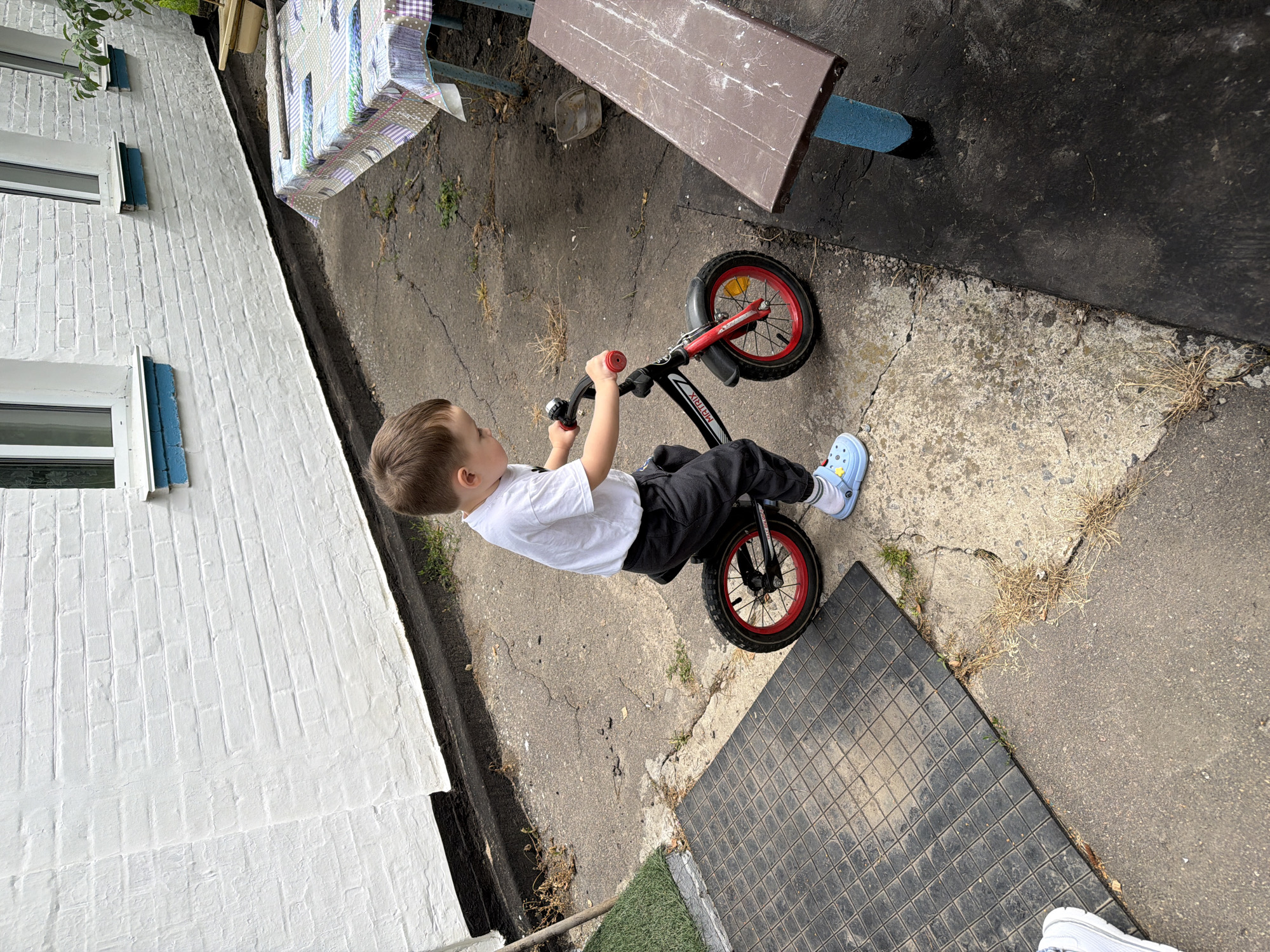 The image depicts a young child, appearing to be around 3-4 years old, on a balance bike in an outdoor setting. The child is wearing a white t-shirt, dark pants, and light blue Crocs with yellow embellishments. The balance bike is black with red rims and appears to be a "Matrix" model. The child is holding onto the handlebars, with their feet on the ground, suggesting they are in the process of riding or learning.
The environment appears to be a paved area, possibly a yard or courtyard. To the left of the child, there is a black mat with a textured surface. In the background, a white brick wall with black trim runs along the side of a building. There are windows with blue trim on the wall. To the right of the child, a picnic table covered with a patterned tablecloth is visible, and further to the right, a wooden bench with blue supports. Dry grass and cracks are visible in the pavement.
There is no specific indication in the image to confirm the location as Kivshovata, Ukraine. The description is based solely on the visual elements present in the photograph.
