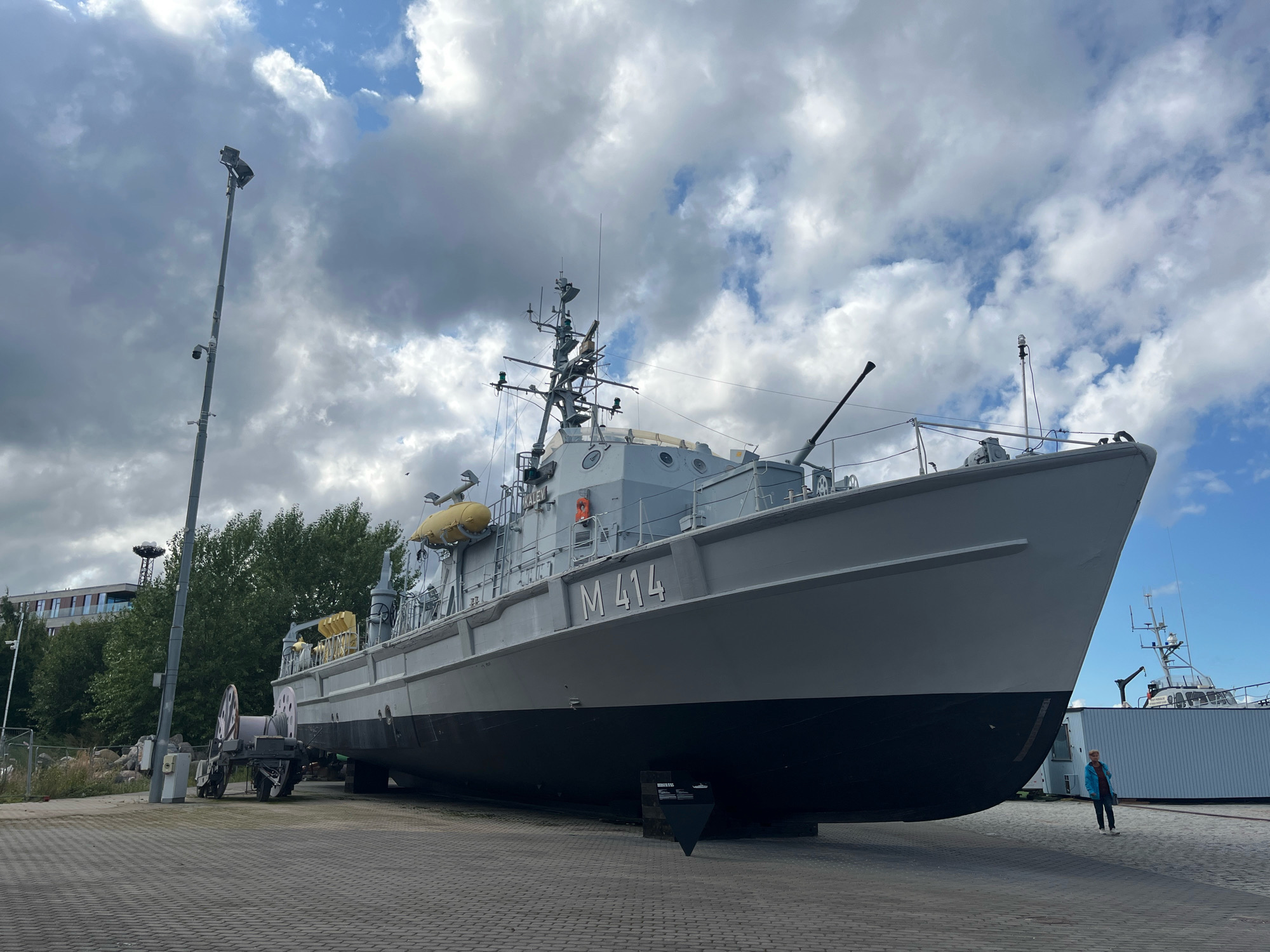 A large, grey, decommissioned patrol boat, marked "M 414," is displayed on a paved area, possibly in a museum or historical site, in Tallinn, Estonia. The boat is positioned on dry land, supported by a visible structure beneath its hull. The boat features several visible armaments, including a cannon mounted on the bow and another on the deck. Yellow life rafts are attached to the port side of the boat. To the left of the patrol boat, a tall, grey pole with lights and security cameras is visible, along with a wheeled cart holding a large coiled rope. In the background, a modern building with multiple windows and trees can be seen. Further to the right, another vessel, possibly a smaller boat, is docked behind a light-colored structure, and a person wearing a blue jacket and dark pants is standing near the bow of the patrol boat, facing away from the camera. The sky is partly cloudy with dramatic formations. The scene appears to be an outdoor exhibit or maritime display.