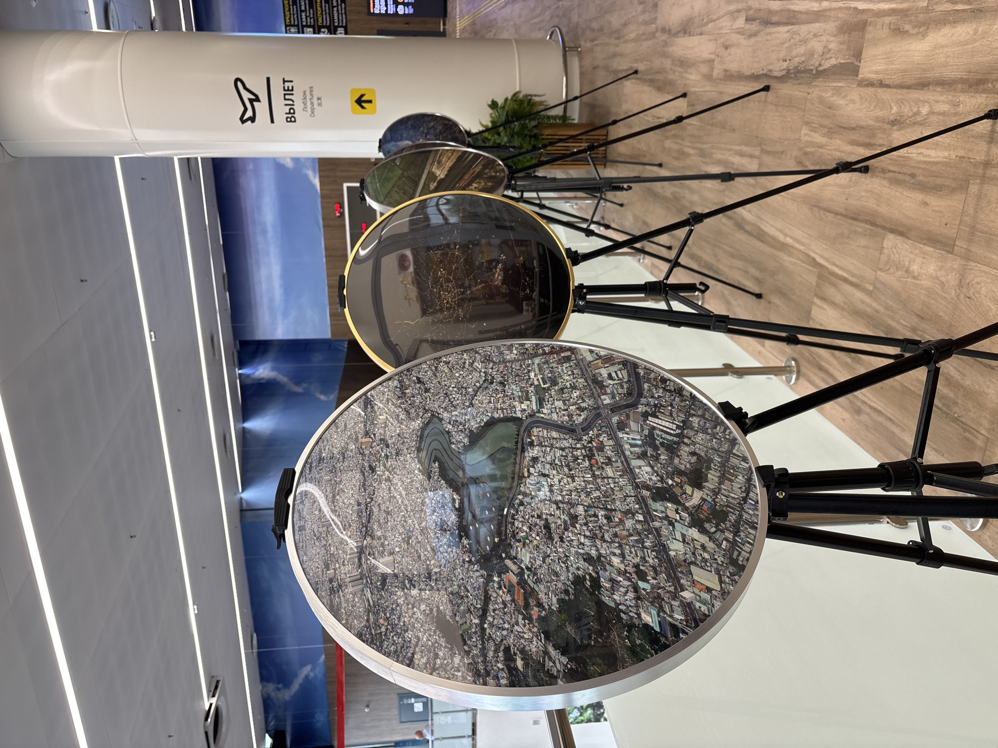 The image shows an art installation in what appears to be an airport or public transit hub, with a display of circular artworks mounted on tripods. The artworks are aerial photographs of urban landscapes. The backdrop is a mural of a sky with clouds, and a white pillar with signage indicating "DEPARTURES" in English, Russian, and Chinese. To the right of the pillar, there is another sign with vertical text that is not legible. The floor is made of light-colored wood planks. The overall environment suggests a modern, clean, and public space. The specific location of Старое Мартьяново, Russia is not indicated by any visual cues in the image.