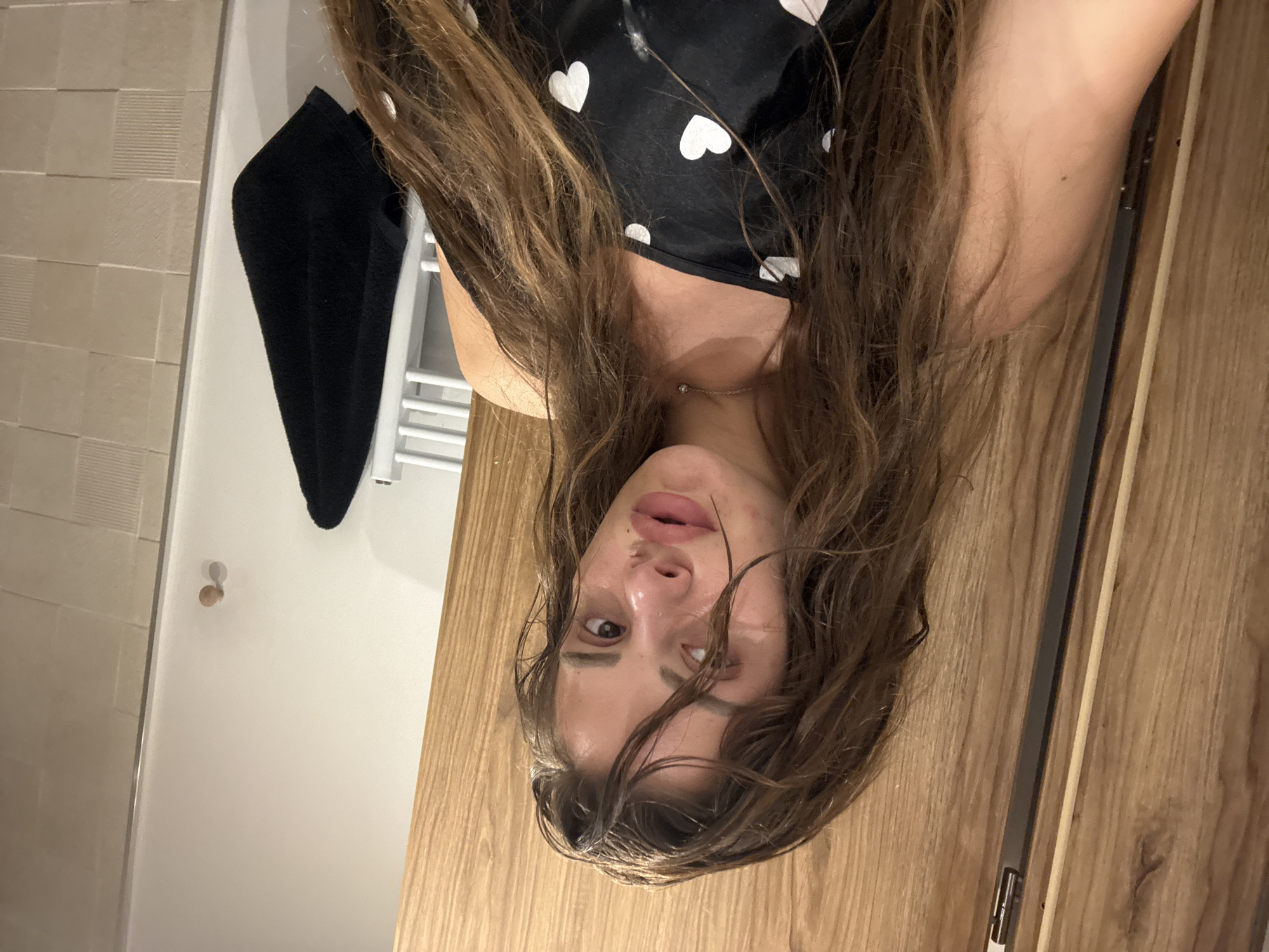 A young woman with long, wet, brown hair is pictured in a selfie. She is wearing a black top with white hearts. She appears to be in a bathroom with wooden cabinets and tiled walls. There is a black towel hanging on a rack in the background.