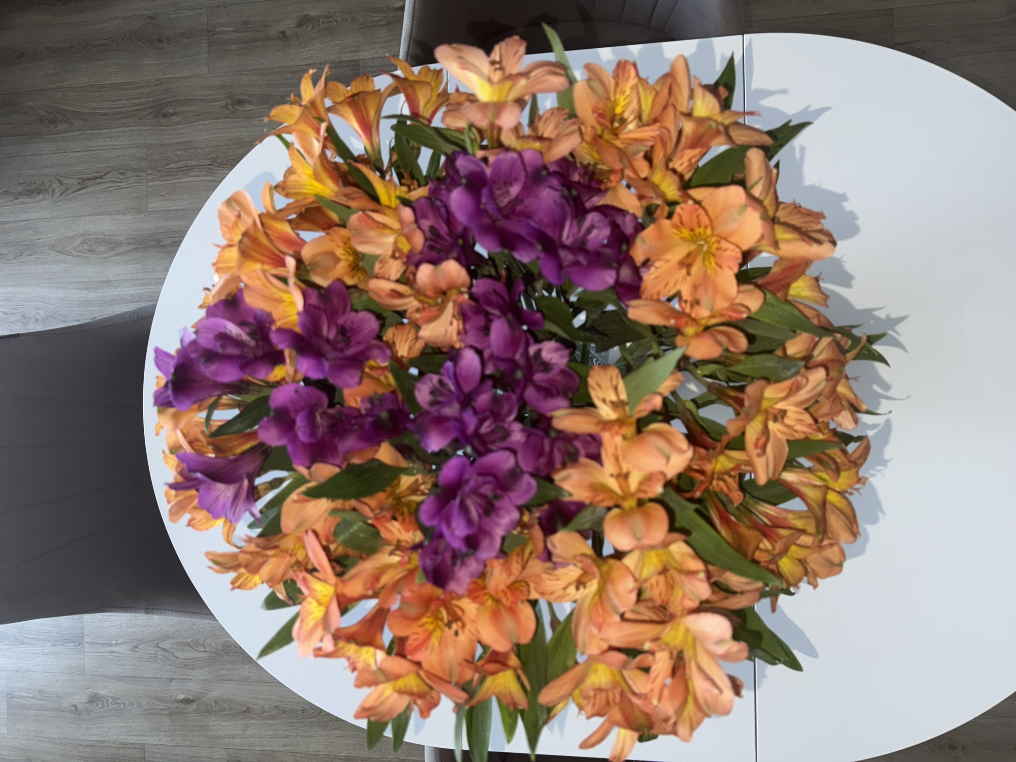 The image shows a floral arrangement on a round, white table. The arrangement includes orange and purple flowers, with green leaves interspersed throughout. The table sits on a grey wooden floor and is surrounded by grey chairs. The location given is the city of Lobanovo, Russia, although there is nothing in the image that would indicate the location.