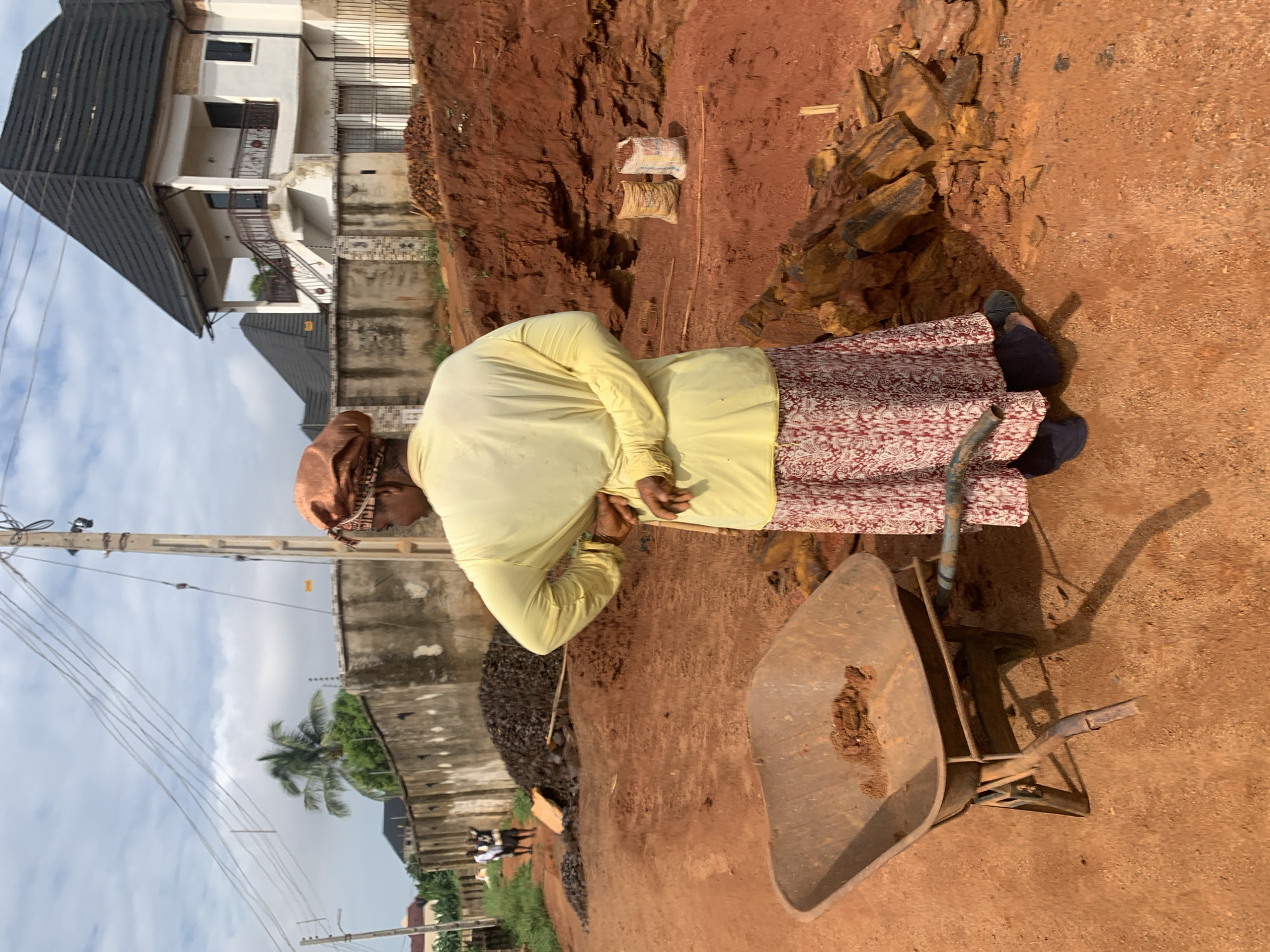 The image depicts a woman in Nsugbe, Nigeria, standing near a wheelbarrow laden with dirt. She is wearing a light yellow long-sleeved shirt and a long skirt with a red and white pattern. She also wears a head covering. She is standing on red dirt, and behind her is a pile of rocks and gravel. In the background, there is a house and a concrete wall. Power lines run along the left side of the image. The sky is visible in the upper left.