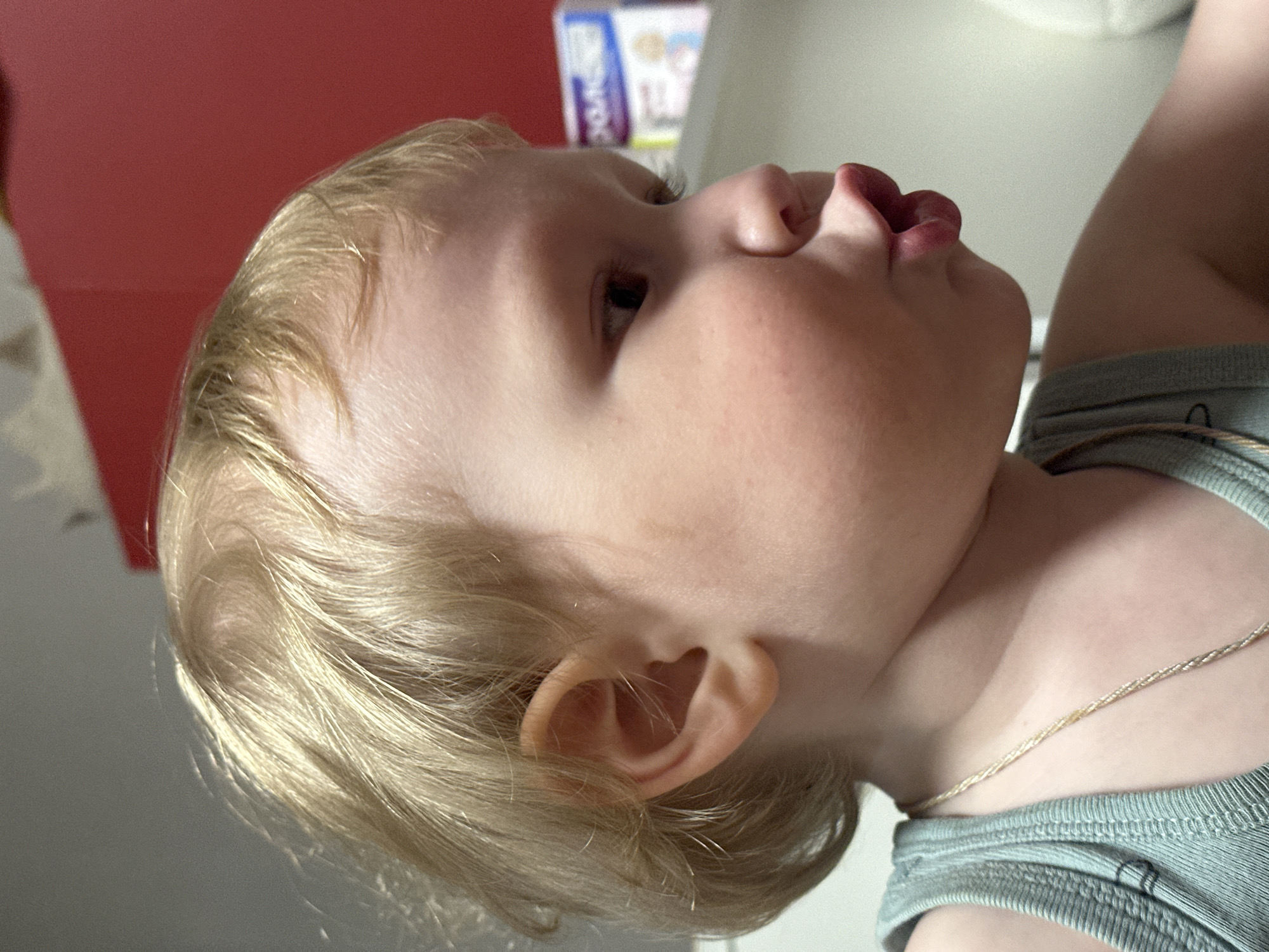 A close-up, tilted shot captures a fair-skinned toddler with blonde hair, light green top, and a gold necklace. The toddler's lips are pursed, as if pouting. The background includes a red surface and a box with indecipherable text. The location is not discernible from the image, so Votkinsk, Russia, cannot be confirmed.