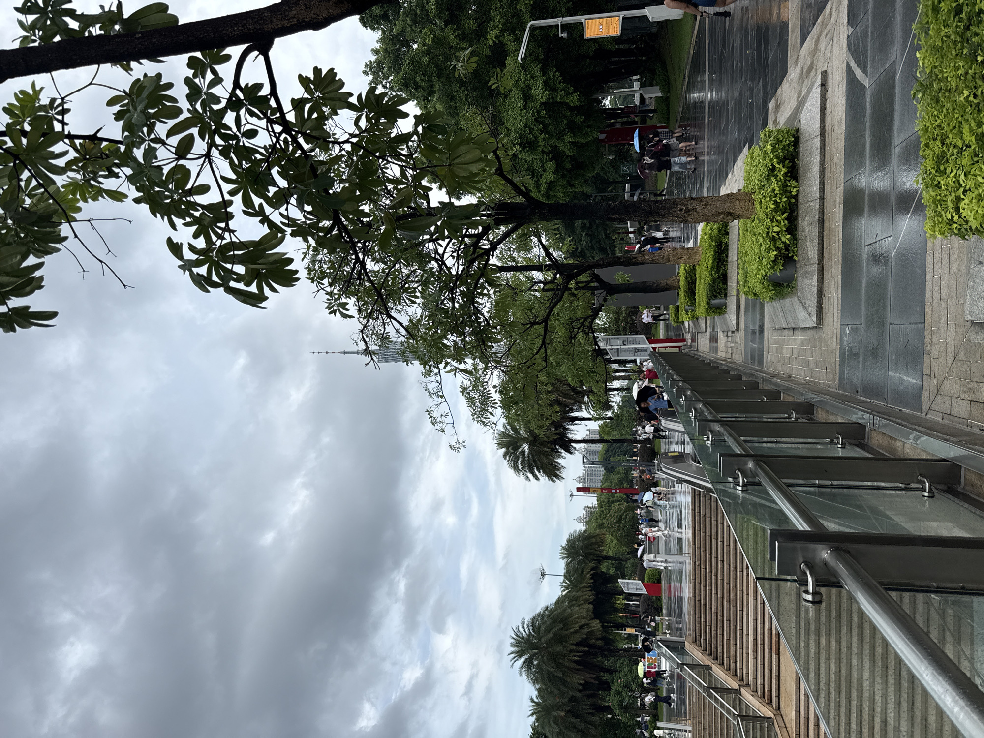 The image depicts a rainy day scene in a park in Tianhe District, China. A paved pathway, wet from the rain, runs diagonally from the foreground to the background. On the right side of the pathway, there are three rectangular planters filled with lush green bushes, each with a tree trunk emerging from the center. Beyond these planters, a dense line of trees forms a verdant backdrop. In the mid-ground, a group of people are walking under umbrellas. Further back, palm trees and other tropical foliage are visible, along with buildings in the distance. To the left of the pathway, a wide set of stone stairs descends, also appearing wet. A glass and metal railing lines the pathway and stairs. The sky is overcast with heavy gray clouds. In the far background, a tall, slender tower, likely a landmark, is visible.
