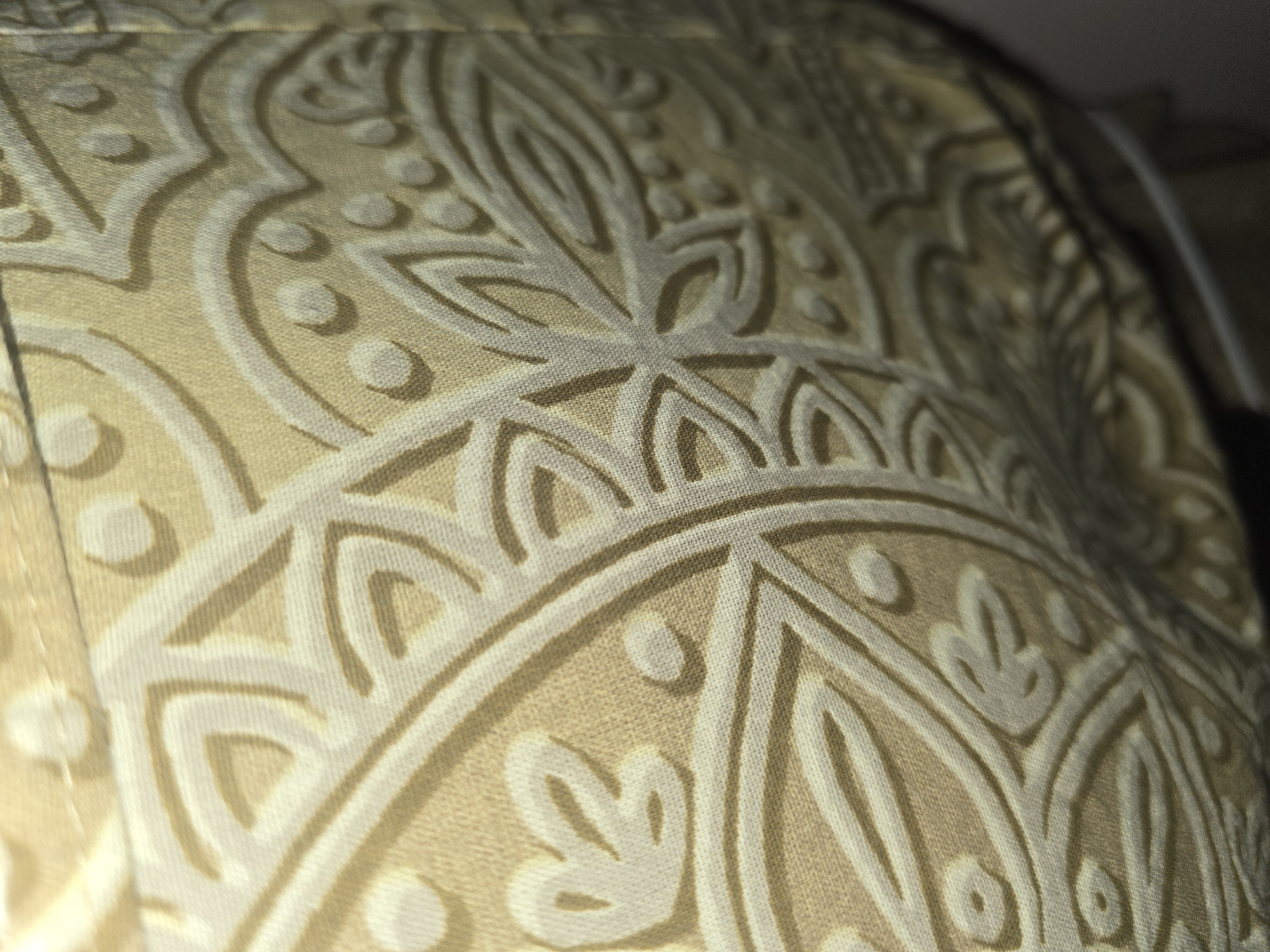The image displays a close-up, angled view of a beige fabric with an embossed, ornate pattern. The pattern features a repeating motif that appears to be floral or paisley-like, with curved lines and circular elements. The lighting creates shadows that highlight the textured nature of the fabric, suggesting it might be a tapestry, upholstery, or a decorative cloth. The background is dark and out of focus, offering no discernible details about the surroundings. There are no people, objects, or actions depicted that would allow for the identification of a specific location within Ulan-Ude, Russia.