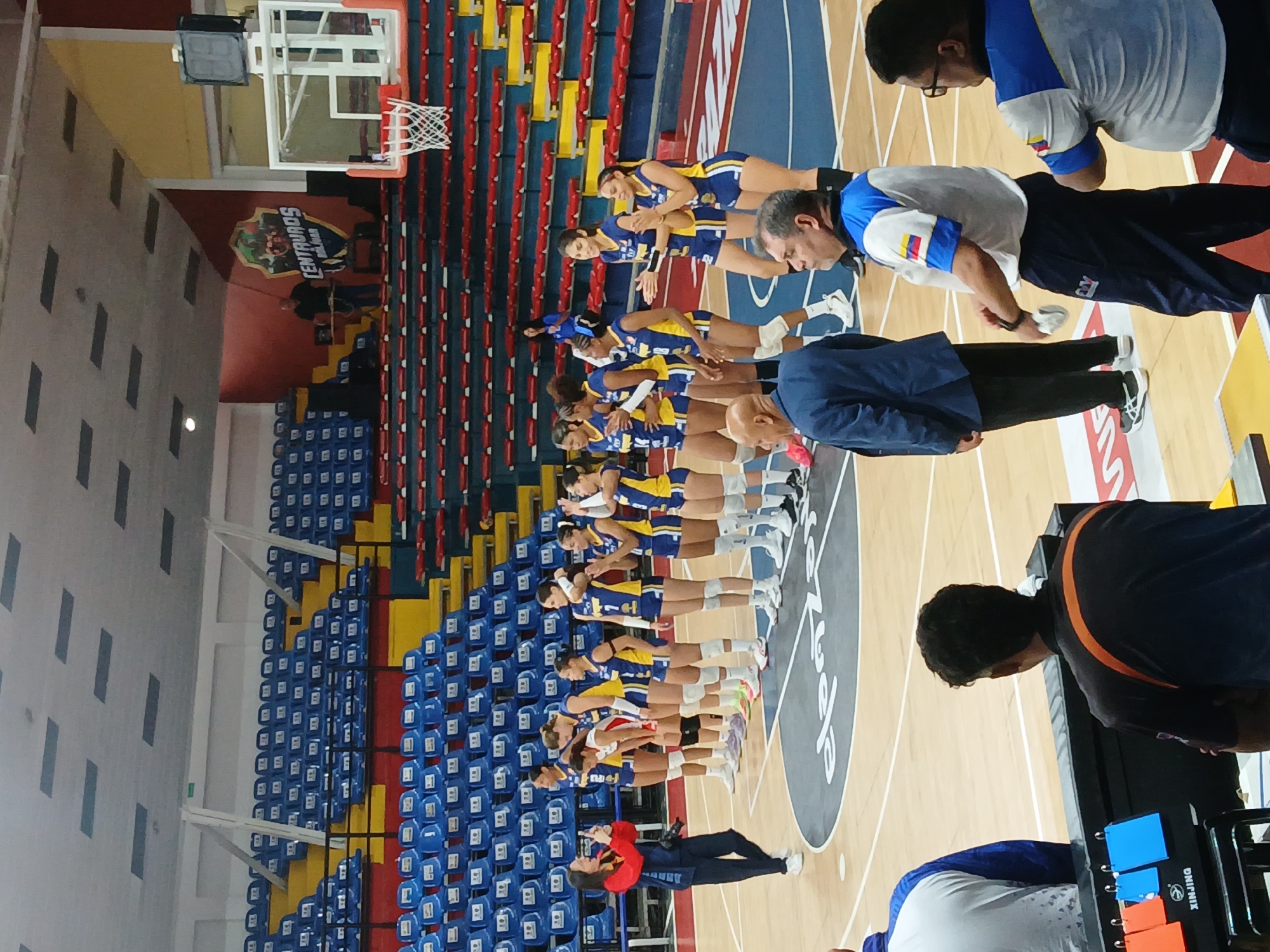 The image shows a women's volleyball team standing in a line on a basketball court inside a sports arena in Ciudad Guayana, Venezuela. The team is wearing blue and yellow uniforms. They are lined up in front of rows of empty blue, yellow, and red stadium seating. Several men are standing near the team, some appearing to be coaching staff, wearing clothing with the Venezuelan flag colors. A basketball hoop is visible in the background, along with a sign for "Centauros Bolivar". The image appears to have been taken at an angle and rotated.