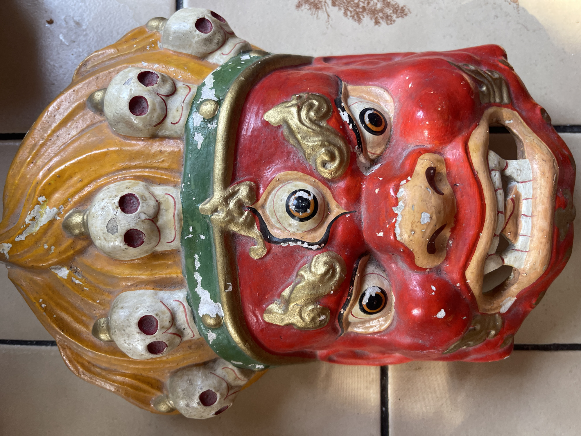 A close-up studio shot depicts a weathered, hand-painted mask featuring a red, demonic face with four eyes, gold accents, and a wide-open mouth revealing rows of teeth. A yellow mane adorned with white skulls lines the left side of the mask, and a green band separates the mane from the face. The mask rests on a surface with tiled flooring. The location is unknown.