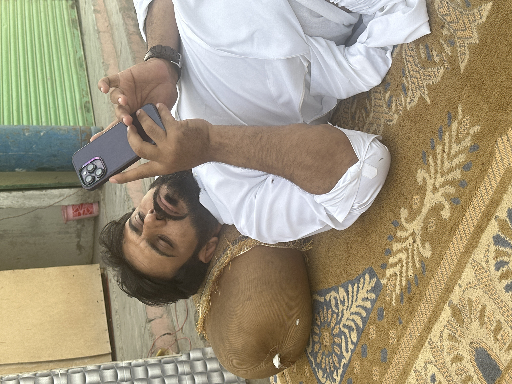A man is lying on a brown and beige patterned rug. He has dark hair and a beard. The man is wearing a white shalwar kameez and a watch on his left wrist. He is holding a dark-colored smartphone in his hands, looking at the screen. His head rests on what appears to be a large, rounded, possibly stuffed object wrapped in brown fabric.
In the background, there is a green corrugated metal surface, a blue cylindrical object, and a red container. The immediate environment around the rug includes what appears to be an outdoor setting with concrete surfaces. The specific location in the city of Unknown, Unknown is not discernible from the image.