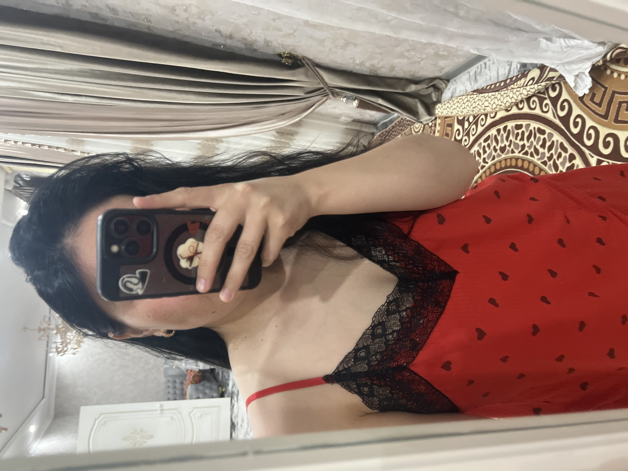 The media file depicts a woman holding a smartphone in front of her face, taking a selfie in a mirror. She has long, dark hair and is wearing a red chemise with black lace trim and a subtle heart pattern. The background suggests an indoor setting, possibly a bedroom, with curtains, a bed with patterned bedding, and furniture visible in the reflection. There are no discernible emotions, time of day, or weather cues. No visible text is present in the image. The location context is Kara-Dıykan, Kyrgyzstan.