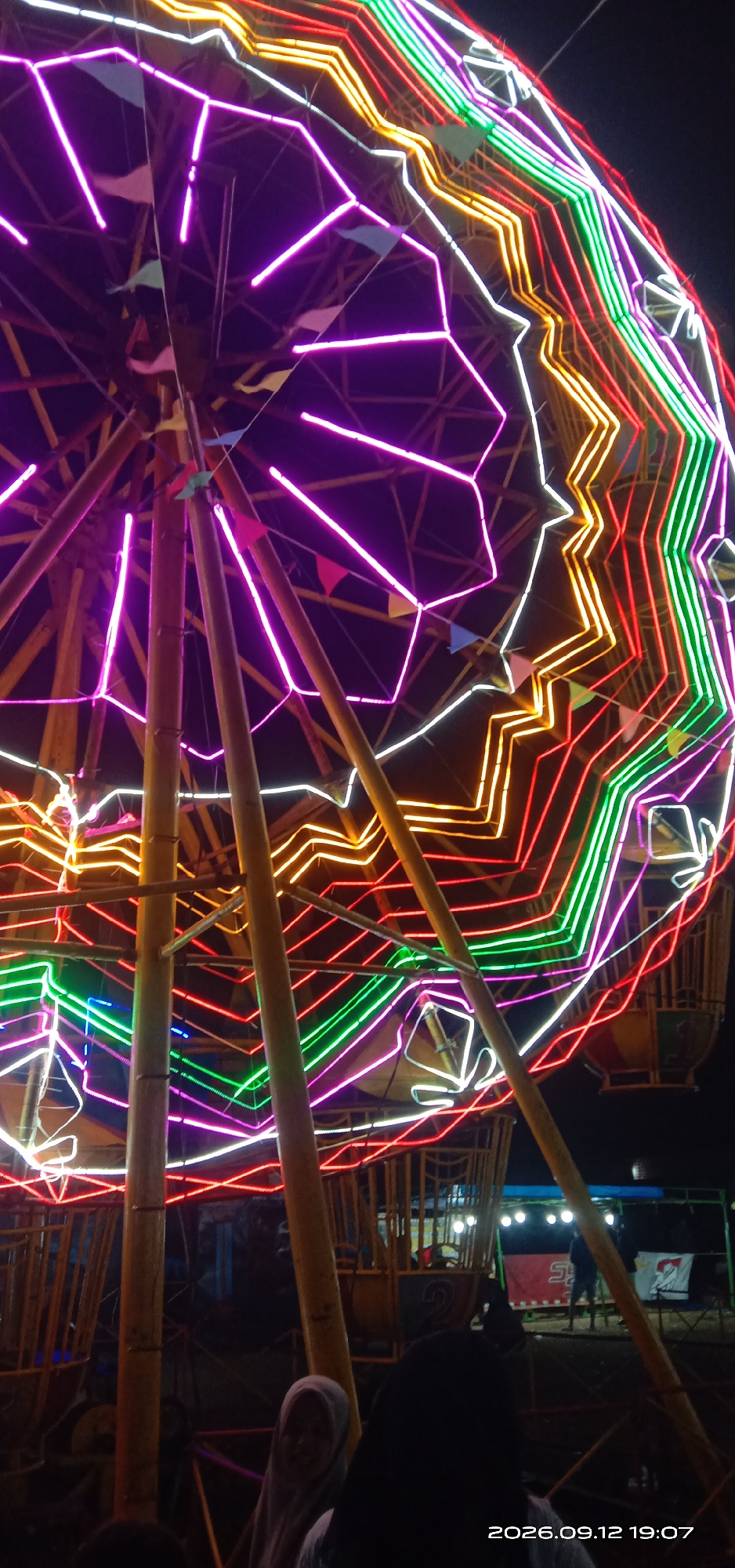 The image shows a brightly lit Ferris wheel at night, likely at a local fair or amusement park. The Ferris wheel is adorned with numerous neon lights in vibrant colors, including purple, pink, white, yellow, red, and green, forming geometric patterns and outlining its structure. Several wooden poles support the central axis of the wheel. Small triangular flags in blue, pink, and green are visible, strung along the structure.
In the foreground and lower portion of the image, several people are present. One individual in the lower right is wearing a light-colored hijab and is partially visible. Other individuals are seen in silhouette, suggesting they are standing or moving around the base of the Ferris wheel.
In the background, beyond the Ferris wheel, there are some structures and additional lights, indicating a larger event or gathering. A banner with red and white colors is visible, along with what appear to be stage lighting. The overall atmosphere suggests a lively evening event. The timestamp at the bottom right indicates the photo was taken on September 12, 2026, at 19:07. The prompt specifies the location as Pakong, Indonesia, which places this scene within that region.