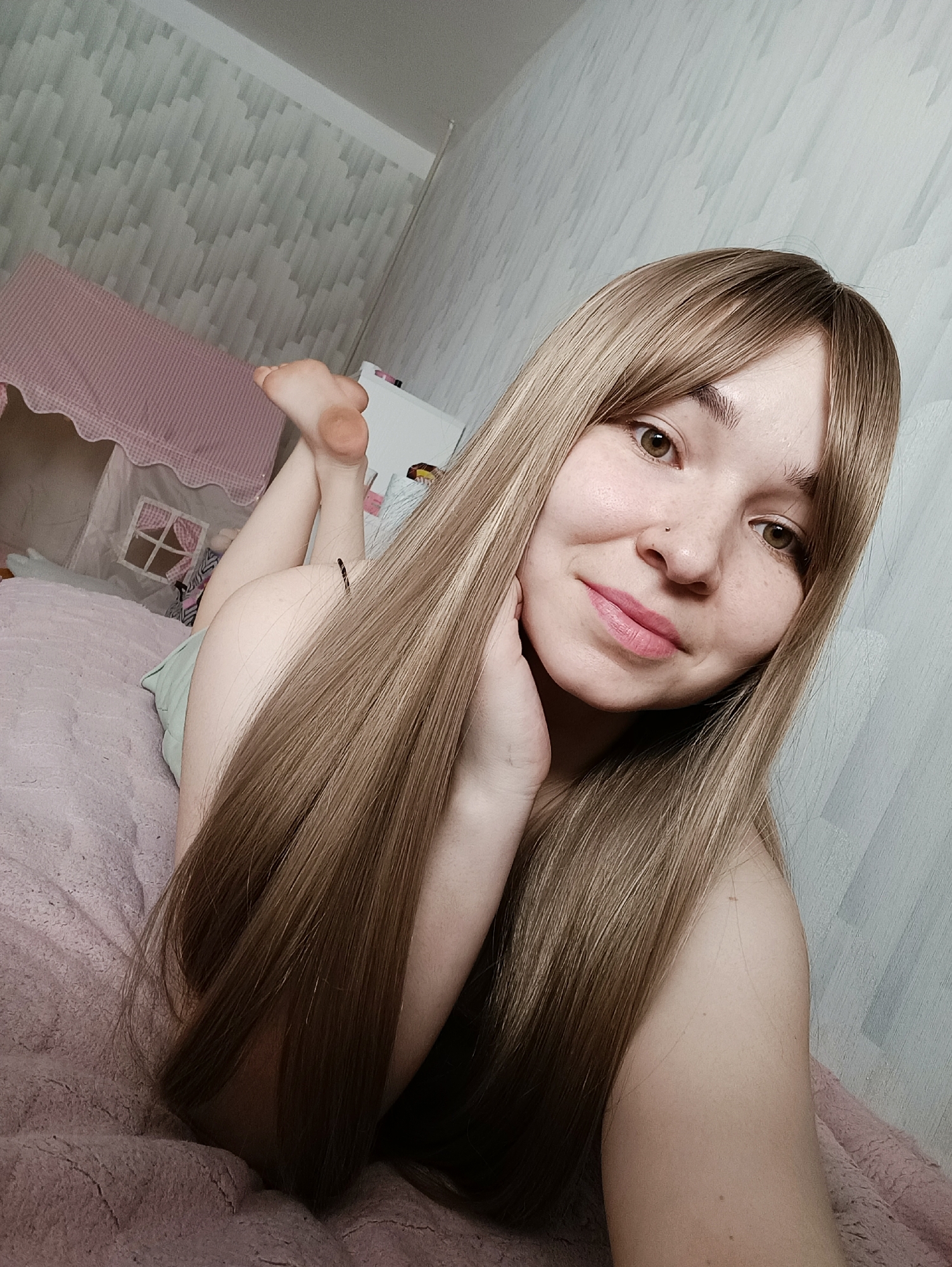 A young woman with long, straight, light brown hair and bangs is lying on her stomach on a pink bed. She is propped up on her elbows, facing the camera with a slight smile. Her right hand is touching her face. Her eyes are brown. She has a nose piercing. Behind her, on the left, is a pink play tent. Further back is a white cabinet. The wall has a geometric pattern. The scene appears to be indoors. There is no information in the image that indicates the city of Izhevsk, Russia.