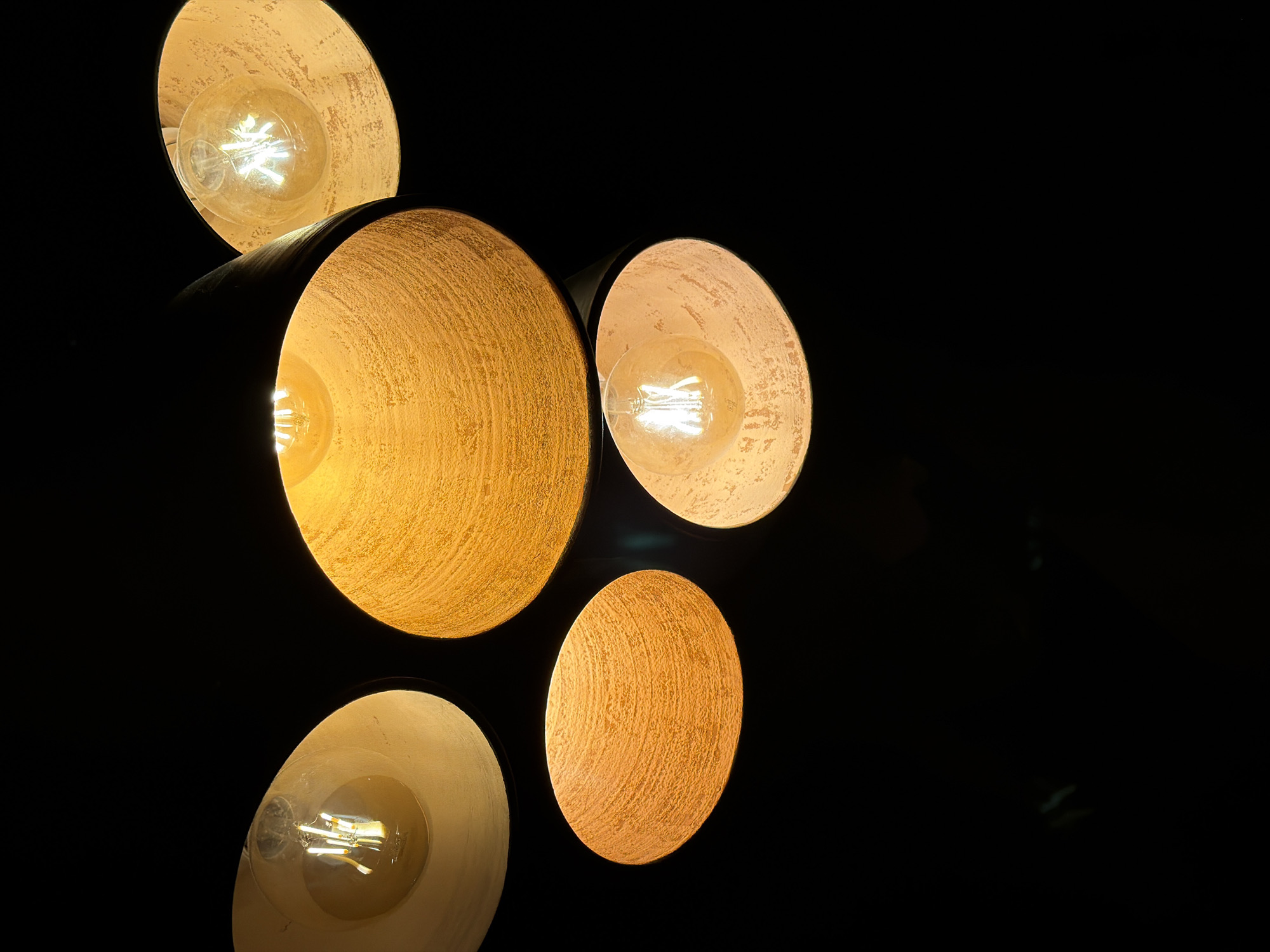 The image features a cluster of five lamps with a dark background. Each lamp consists of a round, bowl-shaped shade, dark on the exterior and textured, light-colored on the interior. The light source inside each lamp is a visible bulb with a glowing filament. The lamps are arranged in a slightly overlapping pattern. No specific location is discernible, and no people, actions, or interactions are visible in the image. The environment is characterized by darkness except for the light emanating from the lamps. The information about Dnipro, Ukraine, is not apparent from the image alone.