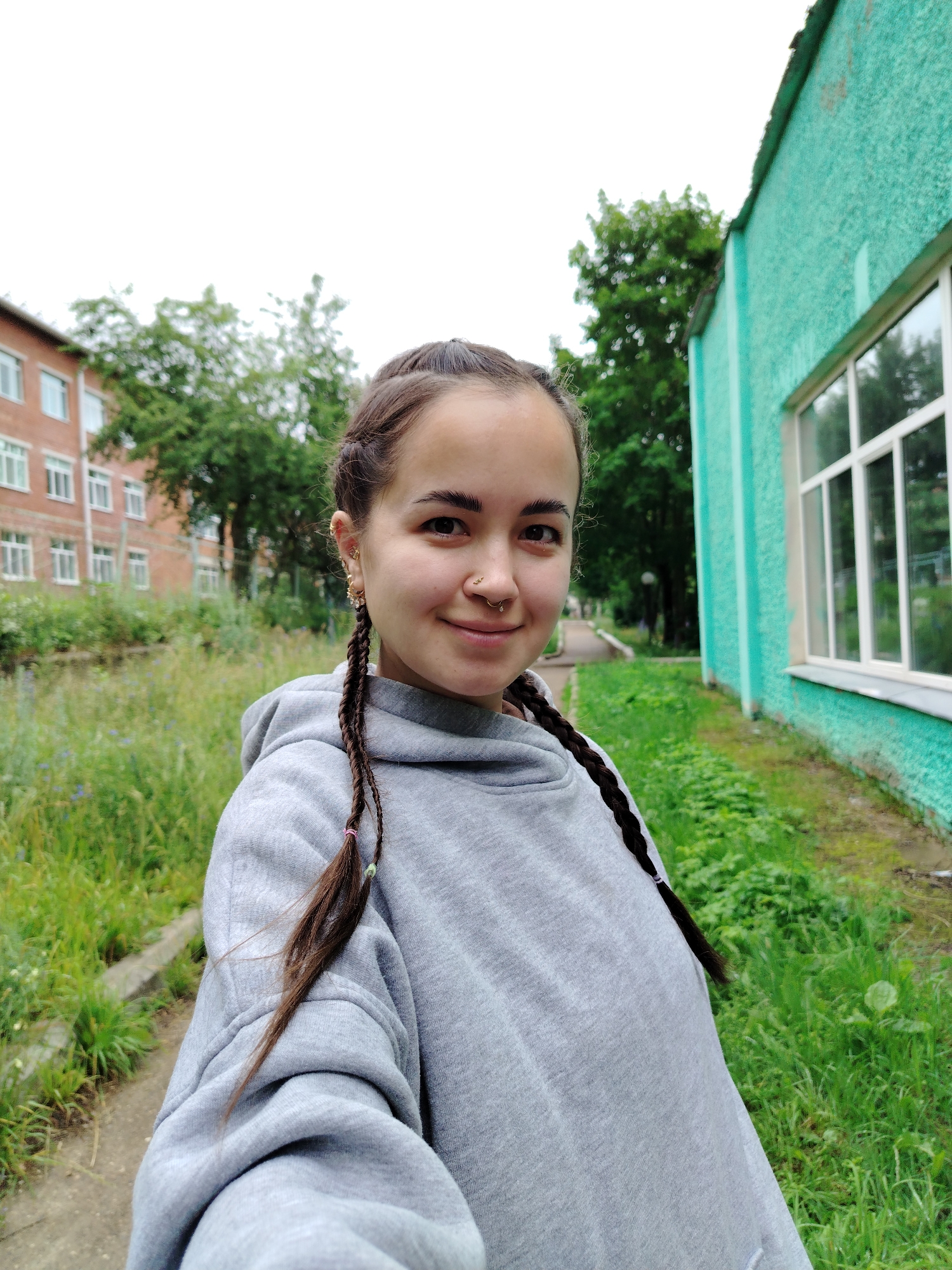 The image is a selfie of a young woman with braided hair and piercings, wearing a grey hoodie. She is standing outdoors in what appears to be a residential area. In the background, there is a red brick building with multiple windows, possibly an apartment complex or school, and a teal-colored building with a large window. Lush green grass and trees surround the area, with a paved walkway visible. The sky is overcast. The scene is consistent with the urban environment of Noviy, Russia.
