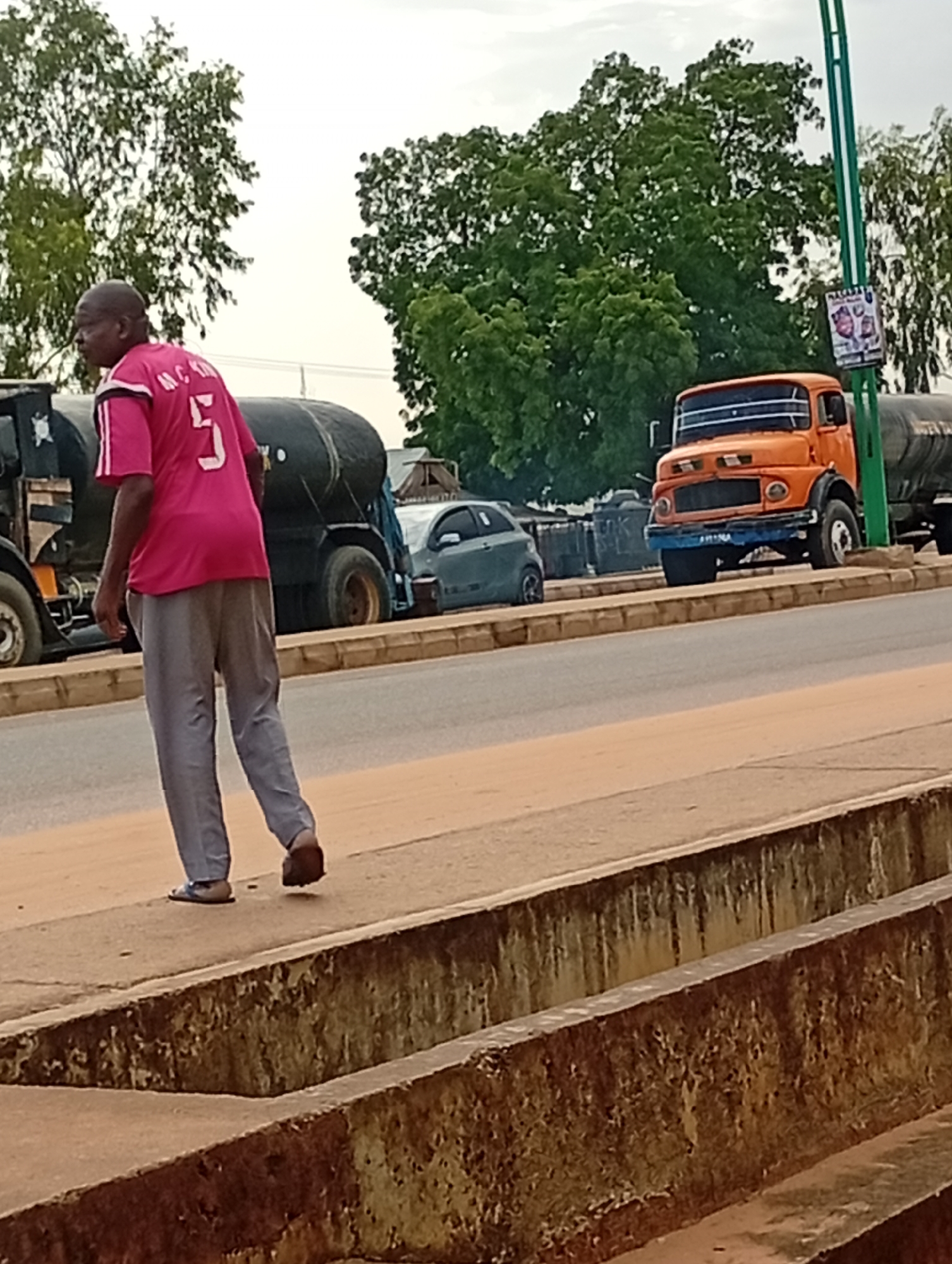 In Tumfure, Nigeria, a bald Black man wearing a bright pink jersey with the number 5 and white trim, and gray pants, walks along a raised concrete walkway beside a road. The man is seen from the back and appears to be walking away from the camera. In the background, several large trucks are parked along the roadside. One truck is an orange tanker truck, and another appears to be a dark-colored truck with a large cylindrical tank. A small gray car is also visible. Trees with green foliage line the background, and the sky is overcast. The scene suggests a typical day in a Nigerian town with ongoing traffic and activity.