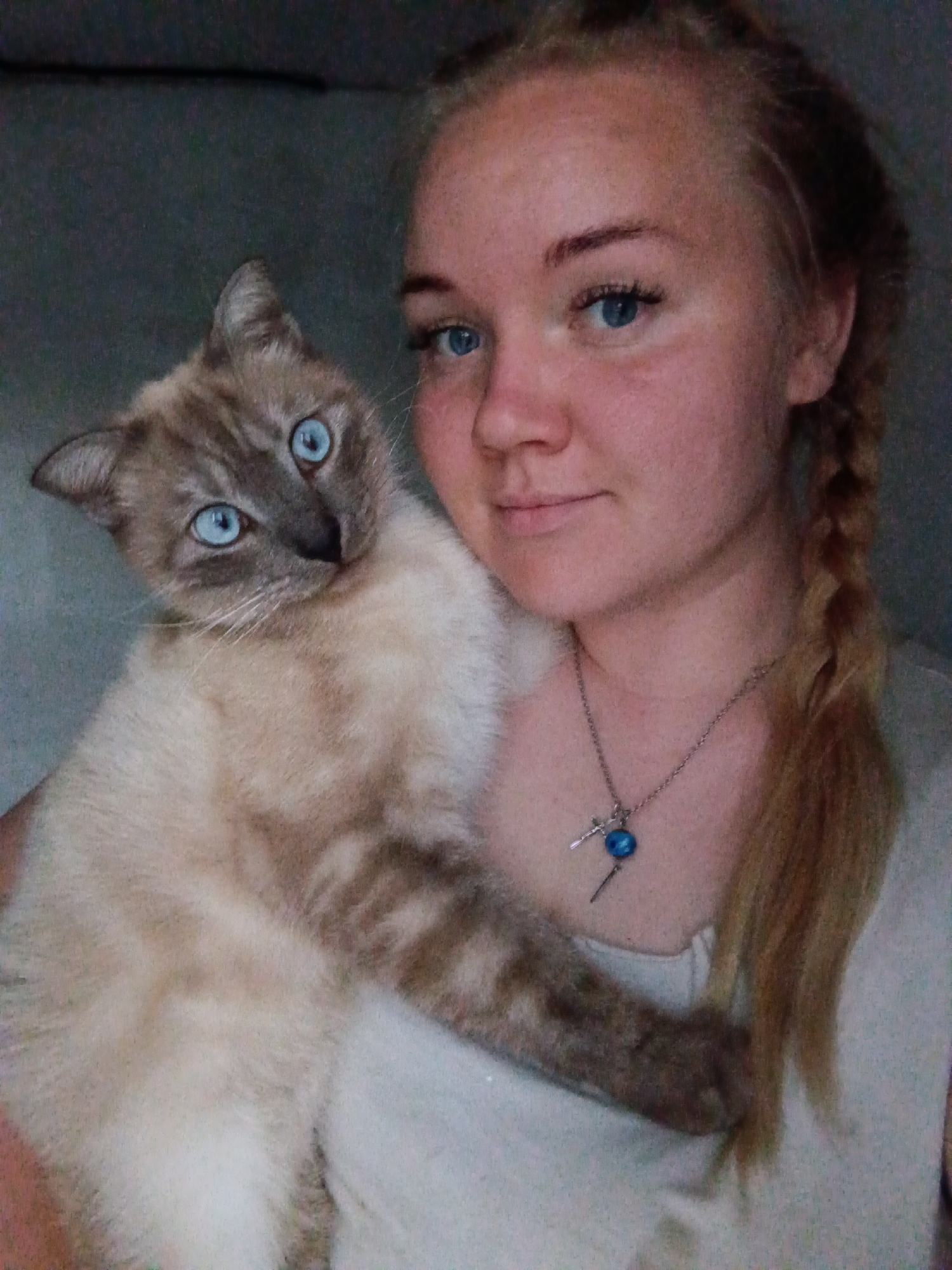 A young, light-skinned woman with blue eyes and blonde hair braided to the side poses with a light brown and white Siamese cat with blue eyes. The woman wears a simple silver necklace with a blue pendant. The background is a plain, muted gray. The city and location of Фоки, Russia, are not determinable from the image.