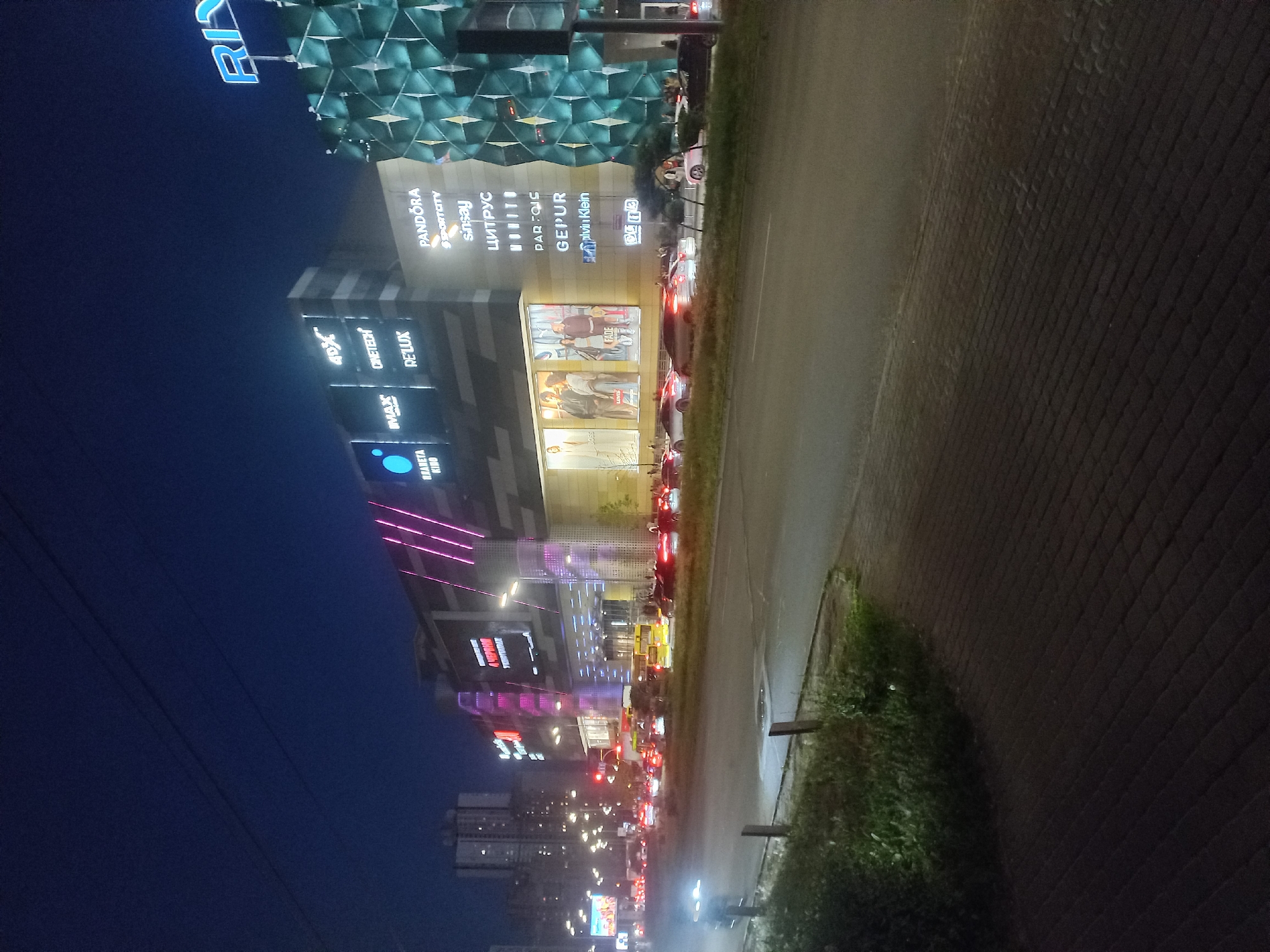 An evening, vertically oriented shot captures a lively urban scene in Kyiv, Ukraine, dominated by a large, brightly illuminated shopping and entertainment complex. The sky is a deep twilight blue, contrasting with the numerous artificial lights that define the cityscape.
On the left, a multi-story building showcases a distinctive, textured facade composed of geometric, scale-like panels in a blue-green hue on its upper section. Below and across several parts of the complex, numerous glowing neon signs are prominently displayed. These include "PANDORA," "SPORTCITY," "sinsay," "ЦИТРУС" (TSYTRUS, a Ukrainian electronics retailer), "PARFOIS," "GEPUR," and a partially visible "Calvin Klein." Further left, signs for a cinema are visible, featuring "ПЛАНЕТА КІНО" (PLANETA KINO, a Ukrainian cinema chain), "MAX," "IMAX," "4DX," "CINETECH," and "RELUX." At the top of the image, a large blue neon sign reads "RI" (likely part of "Respublika Park").
To the right of the buildings, a wide road is visible, appearing wet and highly reflective, mirroring the bright lights from the buildings and traffic. Numerous cars are present, their headlights and taillights creating vibrant streaks of red and white light, indicating active traffic. A sidewalk or paved area runs alongside the road, separated by a narrow strip of grass and dark bushes. The scene conveys the bustling atmosphere of a modern city at night, with commercial activity and vehicular movement.