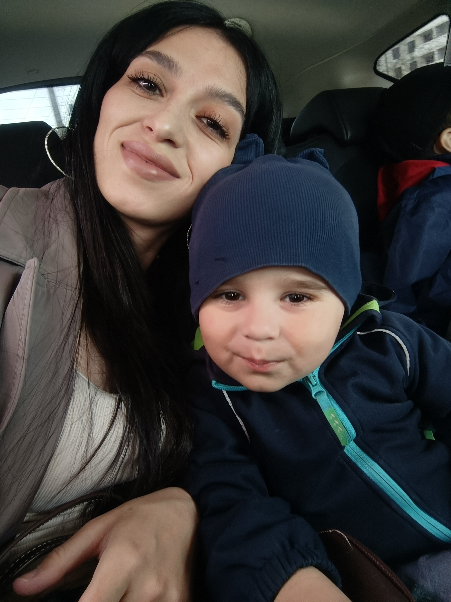 A photograph depicts a woman and a young child in what appears to be the interior of a vehicle. The woman has long, dark hair, fair skin, and is wearing heart-shaped earrings. She is smiling slightly. The child is wearing a dark blue beanie and jacket with a teal zipper and accents. The background shows parts of car seats and a window, suggesting they are inside a car. The photo does not provide enough information to pinpoint the exact location in Molodizhne, Ukraine.