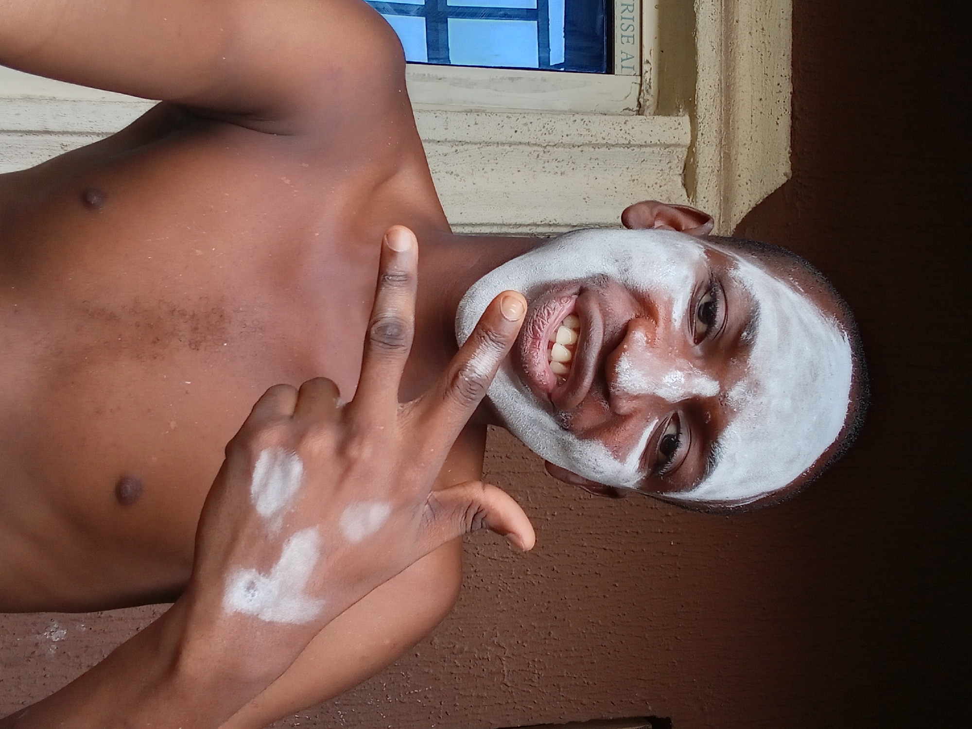 This media file shows a close-up, tilted shot of a young Black person with white cream applied to their face, possibly for a cosmetic treatment or as part of a traditional practice. The person's chest and shoulder are visible, with the skin appearing smooth and dark. A hand is raised towards the face, with white cream also smudged on the fingers and palm. The person is smiling, showing their teeth, and has one eye visible, looking towards the viewer. The background suggests an outdoor or semi-outdoor setting, with a textured wall and a hint of a window with blue panes. The lighting indicates it is daytime. A partial view of text on a dark blue object in the background appears to read "RISE AT". The overall mood seems casual and playful. The context provided indicates this scene is from Nkpor, Nigeria.
