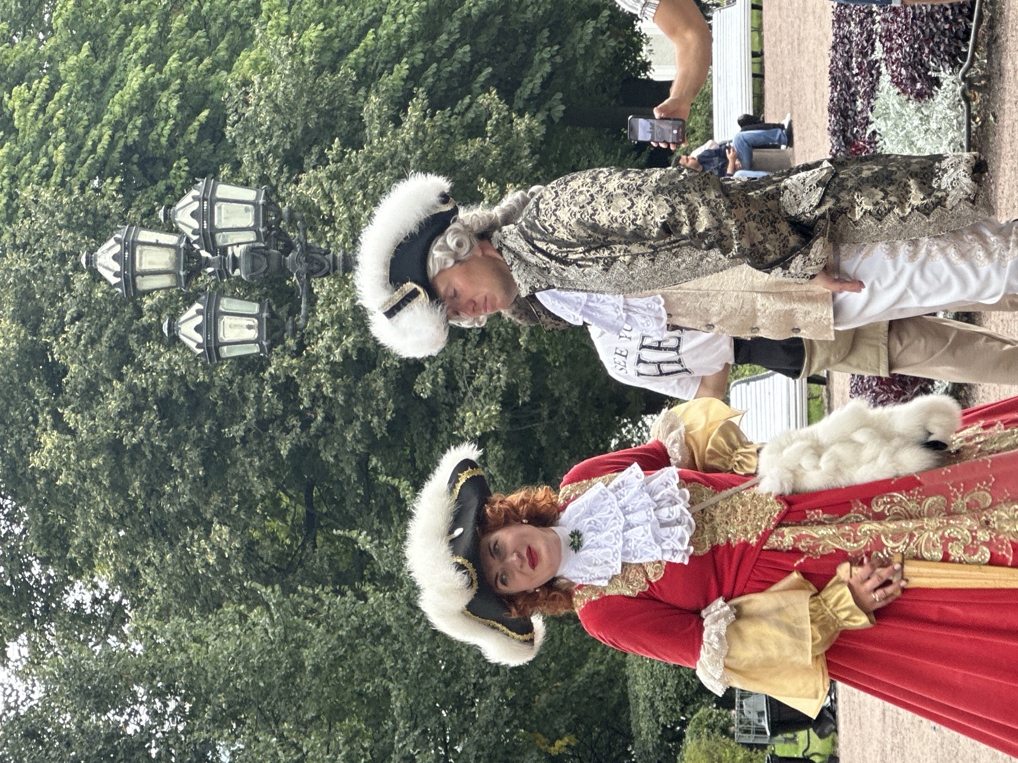 A full-shot medium of two people dressed in 18th-century attire standing in front of lush green trees, with a three-headed streetlamp behind them. The person on the left is a woman with red curly hair wearing a large black hat adorned with white feathers, a red gown with gold trim, and a ruffled white jabot. She is also carrying a white fur muff. The person on the right is a man with a powdered wig, also wearing a black hat with white feathers, a gold patterned coat, and white pants. His gaze is directed to the left, while the woman's is directed forward. In the background, several people are seated on benches, and one person is holding up a smartphone, possibly taking a photo. The ground appears to be paved with gravel. The overall impression is that of a historical reenactment or a themed event taking place in a park. The iconic lamppost and trees suggest a location like Peterhof Palace or Catherine Palace, both famous for their elaborate gardens and historical significance within the city of Saint Petersburg, Russia.