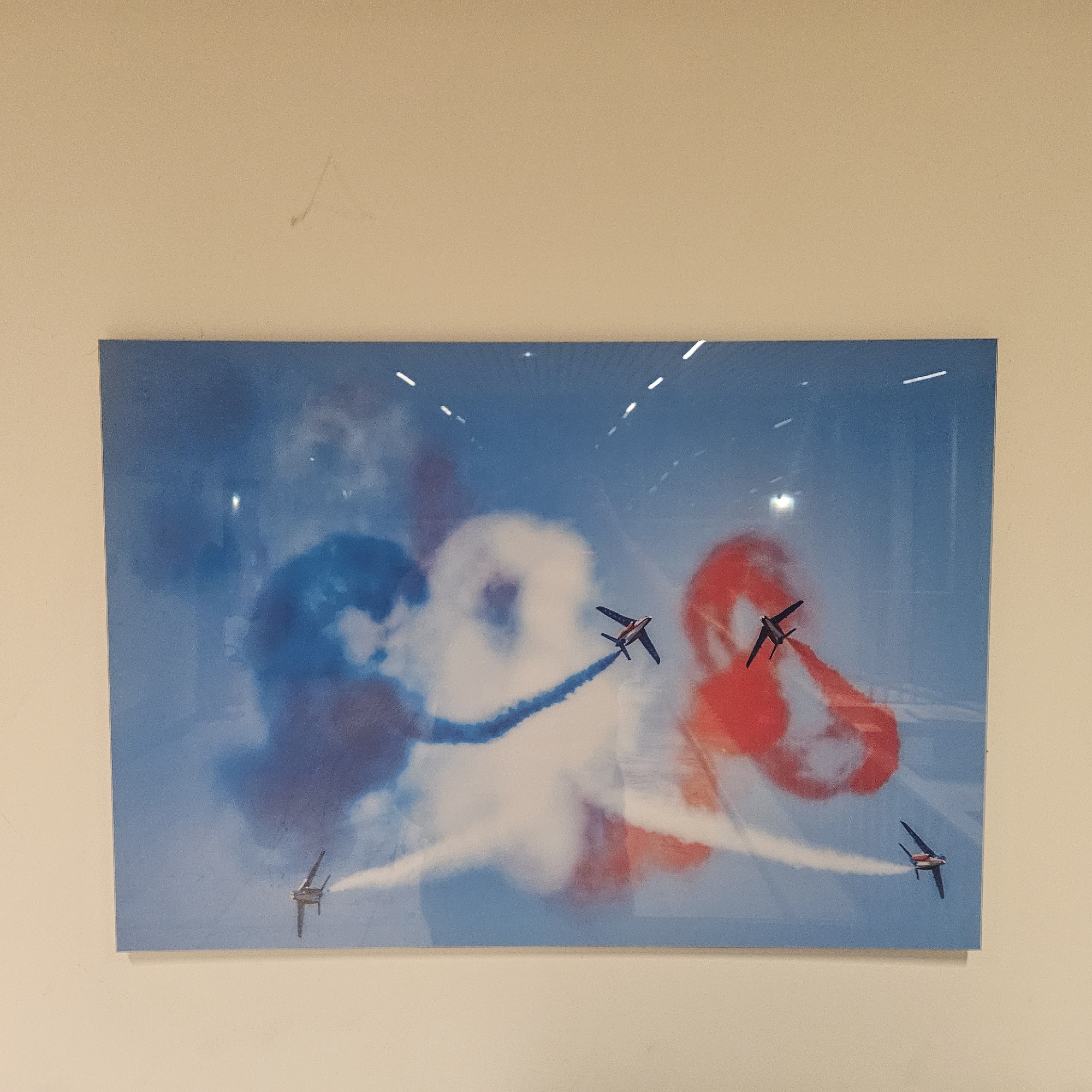 The image is a photograph of a framed artwork depicting a skydiving or airshow event. The artwork features a blue sky with clouds in red, white, and blue, reminiscent of the French flag. Several small planes are visible, trailing smoke in the colors of the French flag. The artwork is displayed on a light beige wall. There are no people visible in the image. The location of this artwork in Lançon-Provence, France is not discernible from the image.